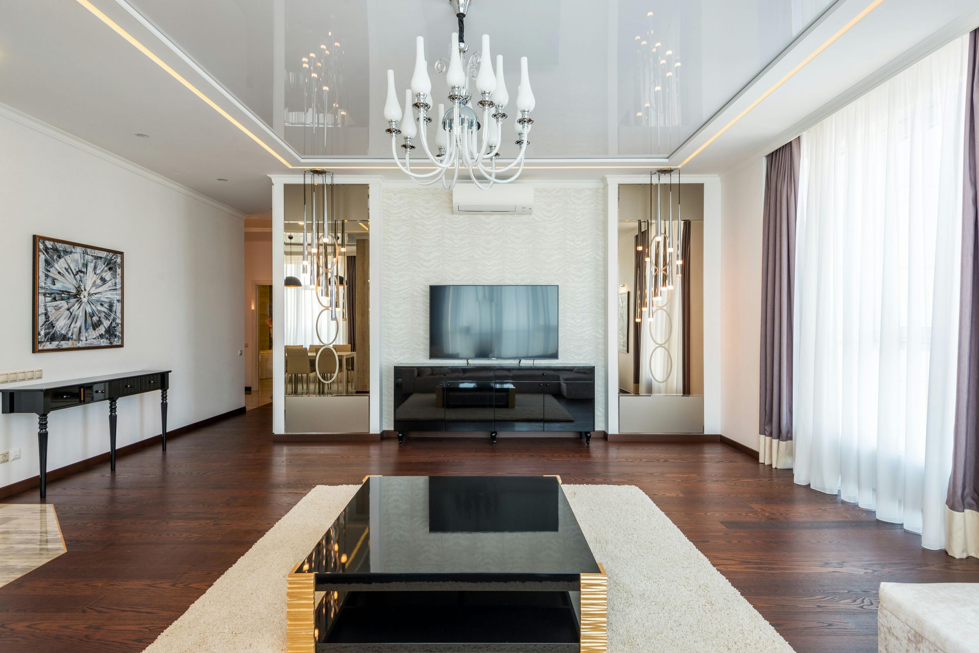 A modern living room with dark wood floors, a black coffee table on a white rug, a large TV, and a glass chandelier.