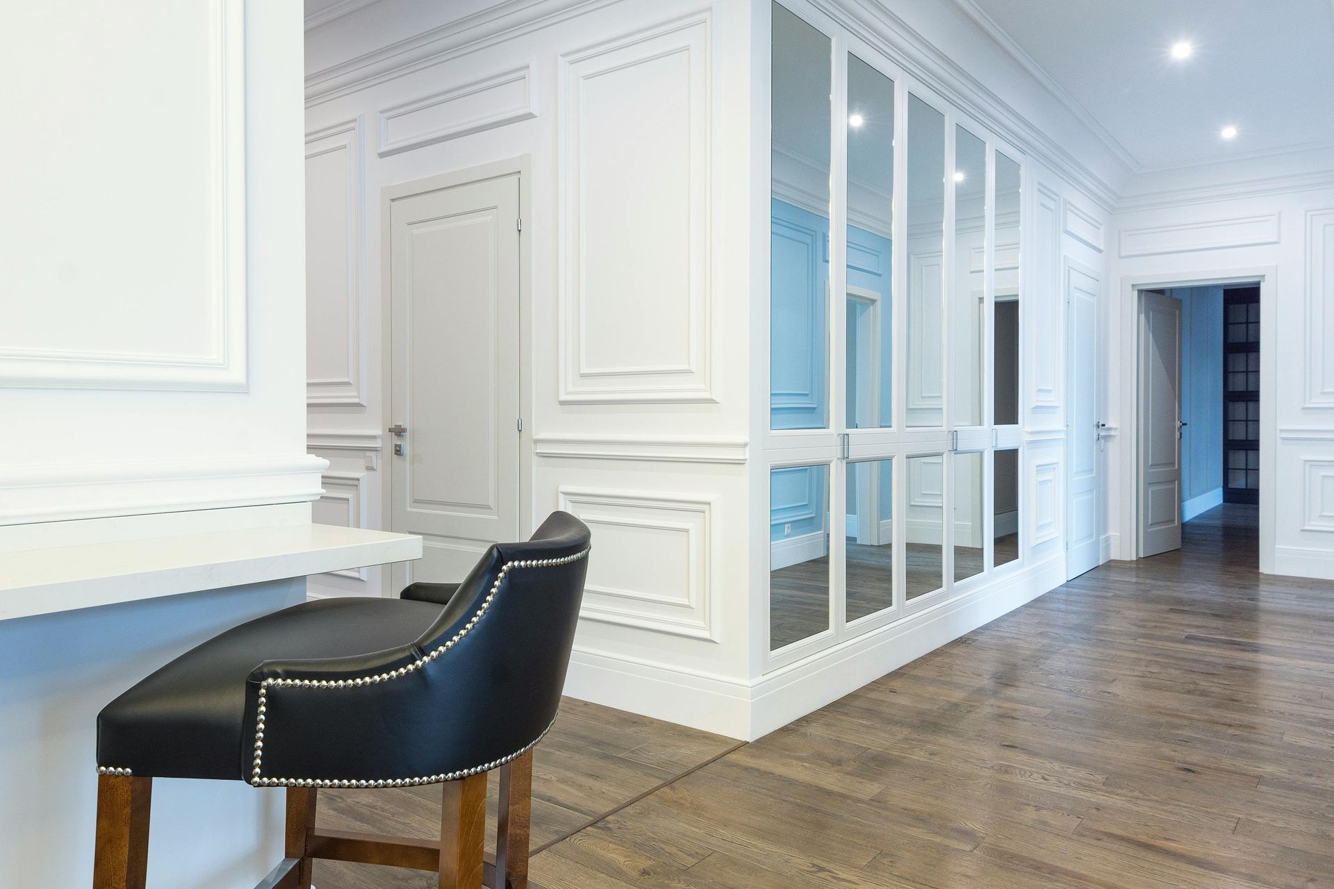 A high-angle view of a modern hallway with white panelled walls, wooden flooring, a black stool, and mirrored cabinets.