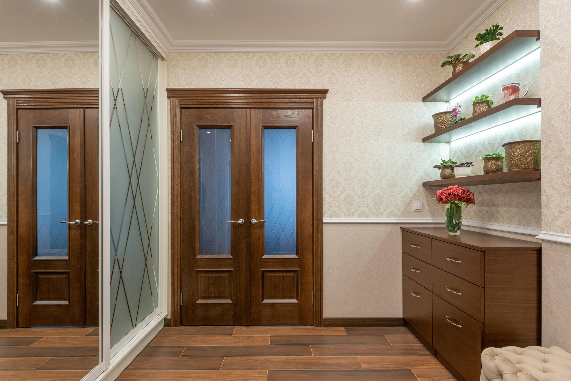 Modern entryway with dark wood doors, a mirrored closet, wooden dresser, and floating shelves with plants on patterned wallpaper.