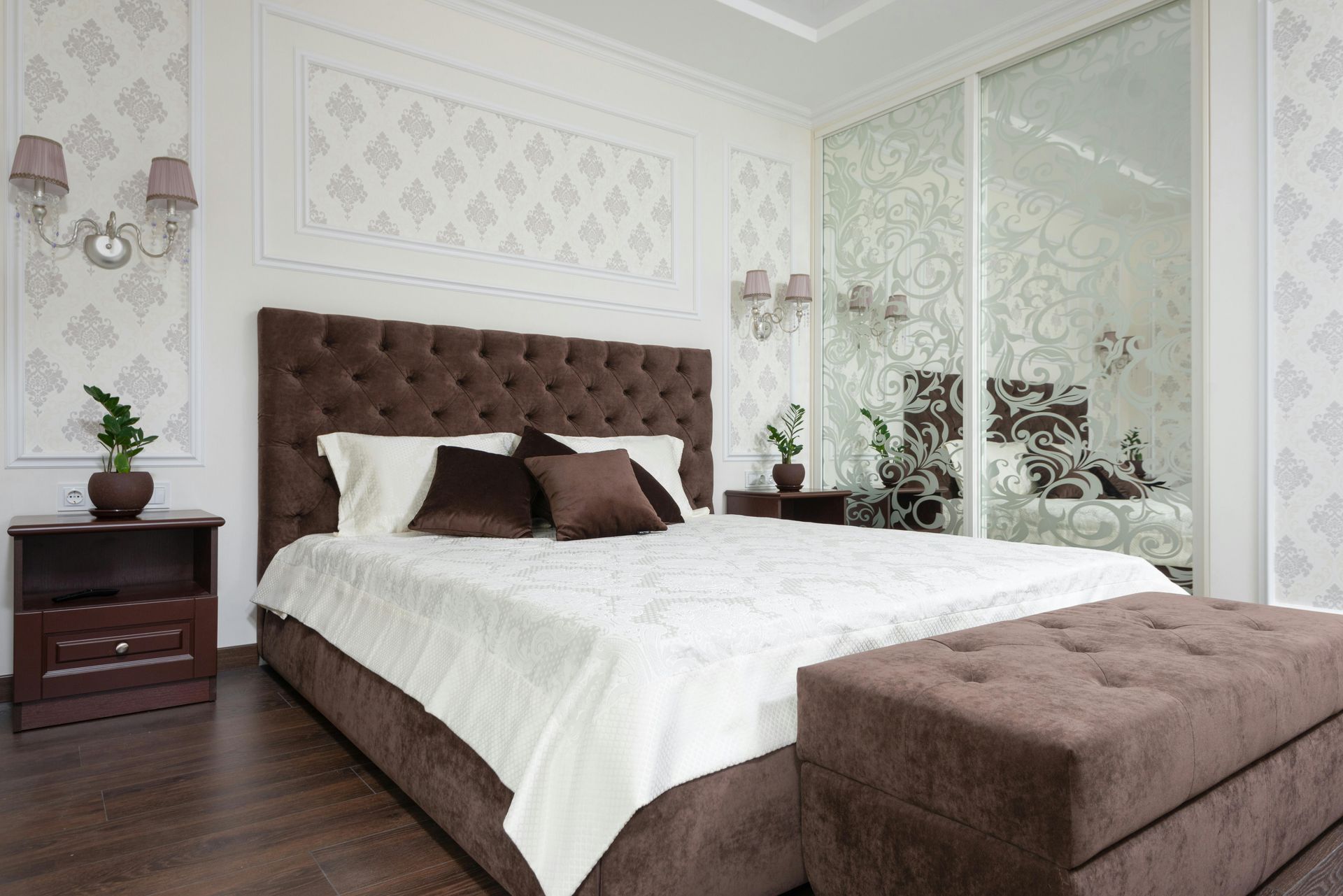 A modern bedroom featuring a tufted brown headboard and matching bench, white bedding, and mirrored closet doors.