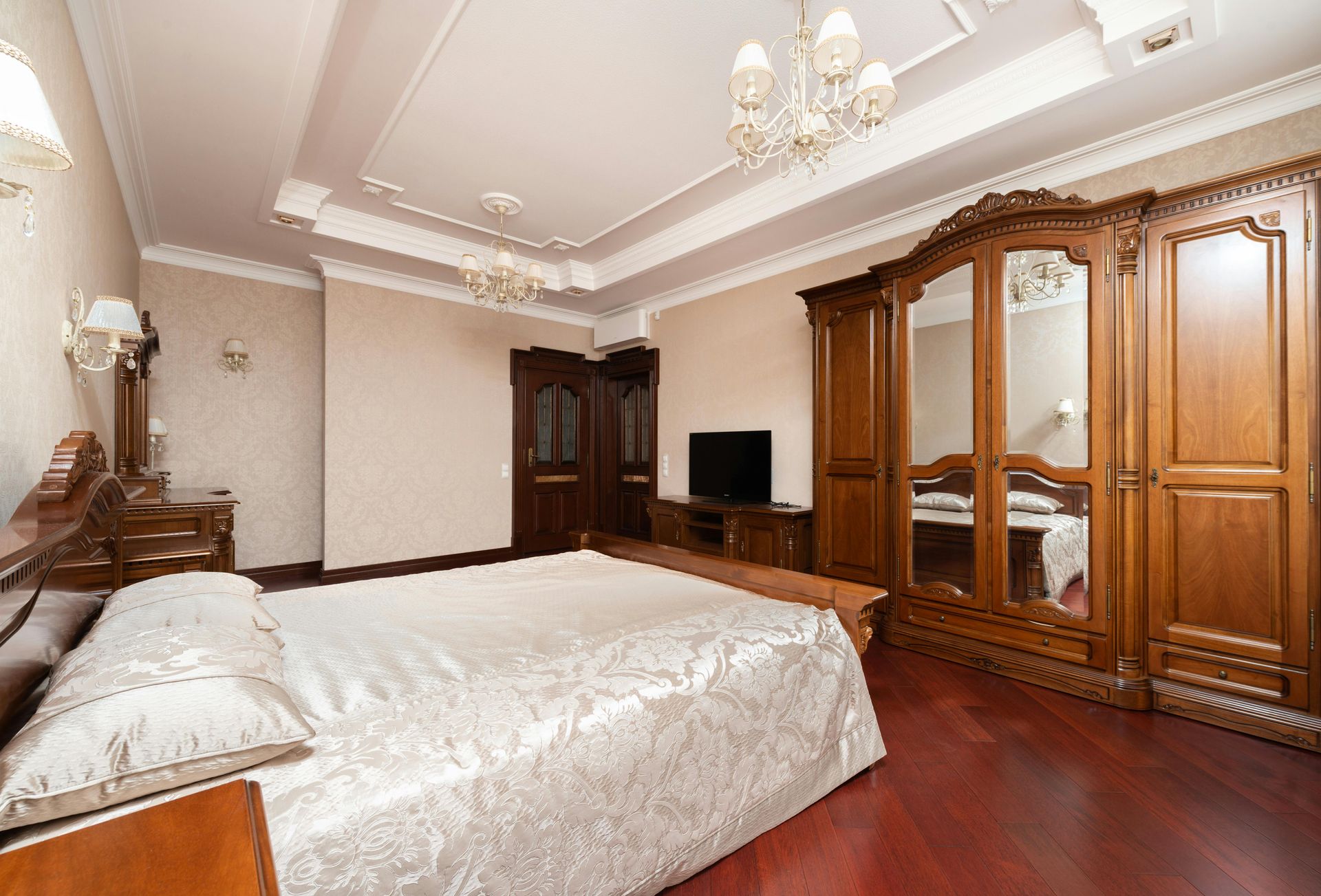 A bedroom with a bed featuring a textured beige bedspread, a large wooden wardrobe with mirror doors, and a television.