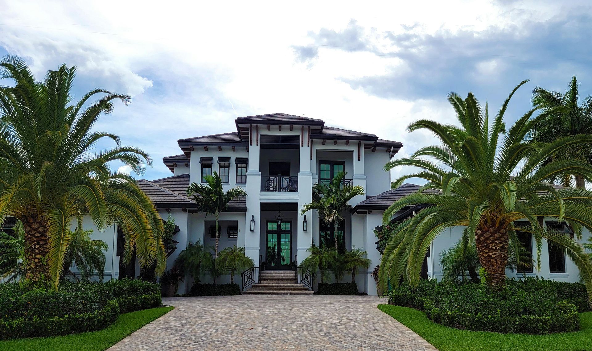 A two-story white luxury house with a stone driveway and palm trees in a front yard under a cloudy sky.