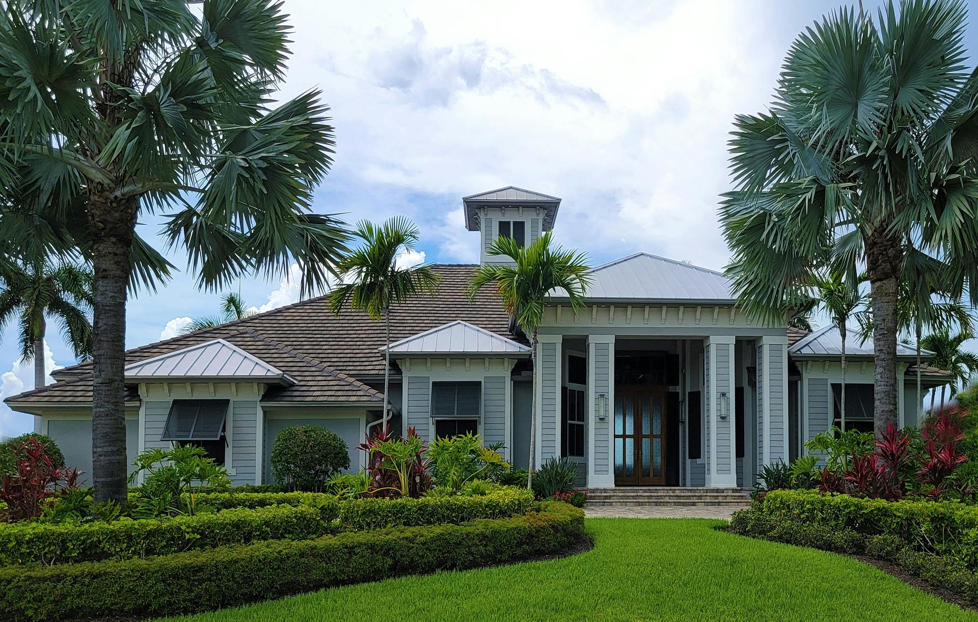A light-colored house with a tiled roof surrounded by manicured hedges and tall palm trees under a cloudy sky.
