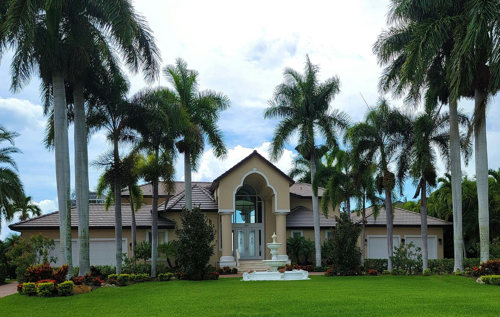 A tan-colored suburban home with a central arched entrance, surrounded by tall palm trees and a green lawn.