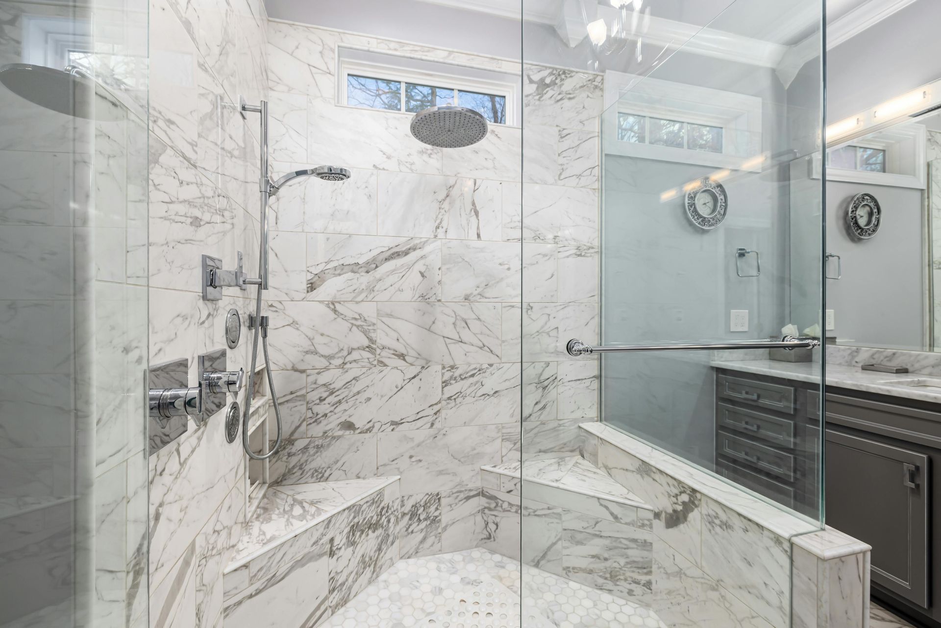 A luxurious marble shower with built-in corner benches, a rainfall showerhead, and a glass door, adjacent to a vanity.