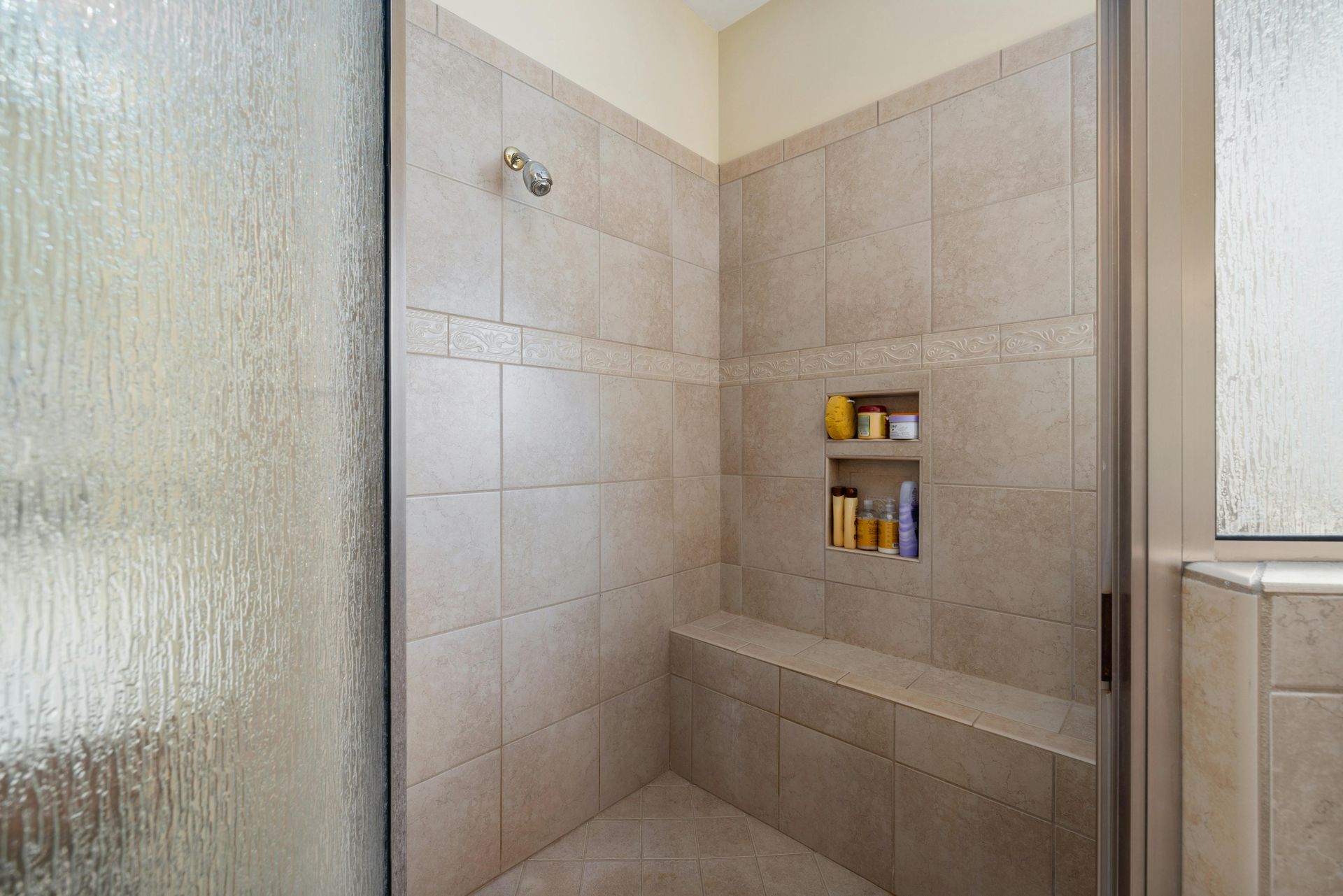 A tiled walk-in shower with a built-in bench, a shower head, a two-tiered wall niche with toiletries, and frosted doors.