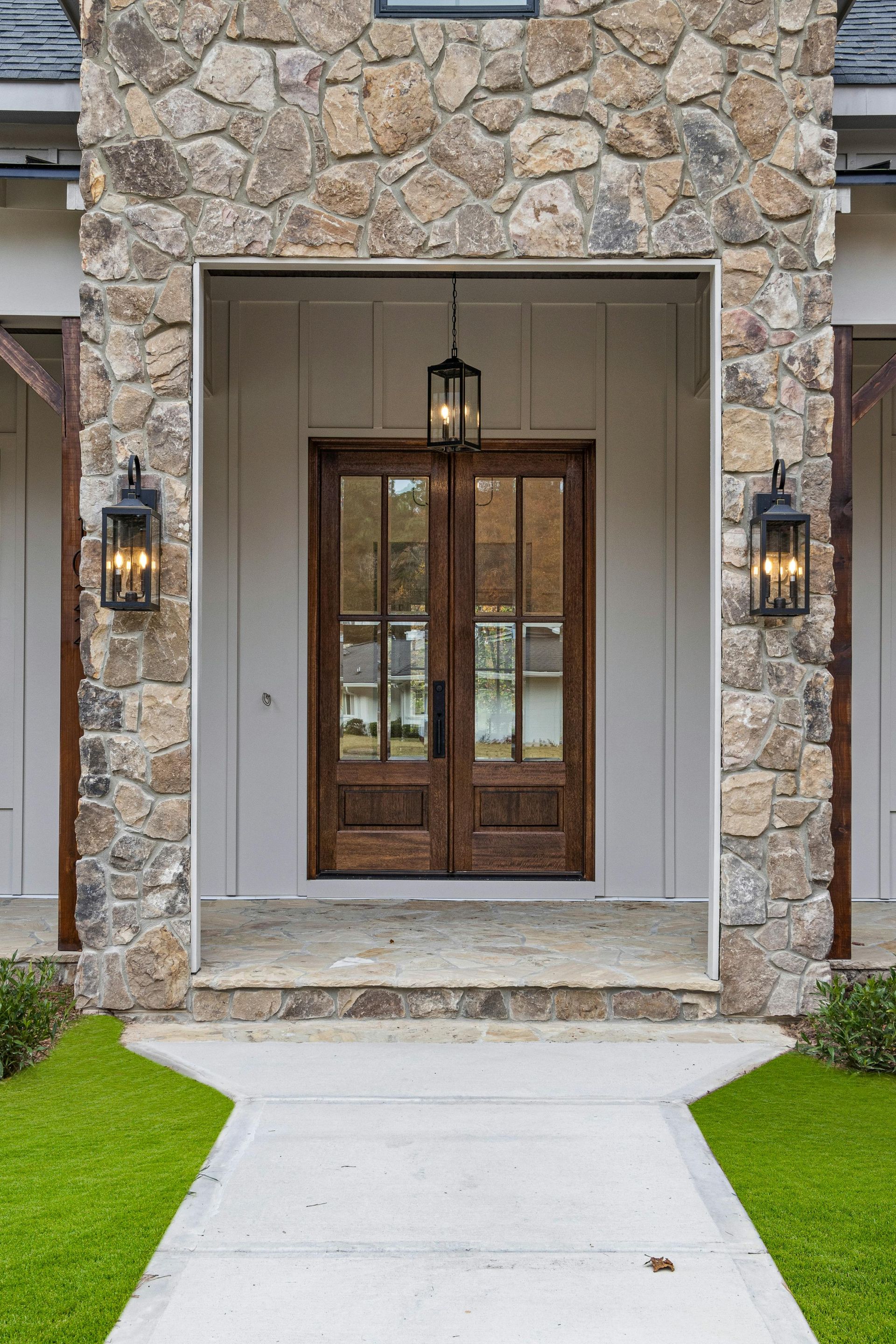 A modern home entrance with a wood double door, white vertical siding, stone masonry, and three black lanterns.