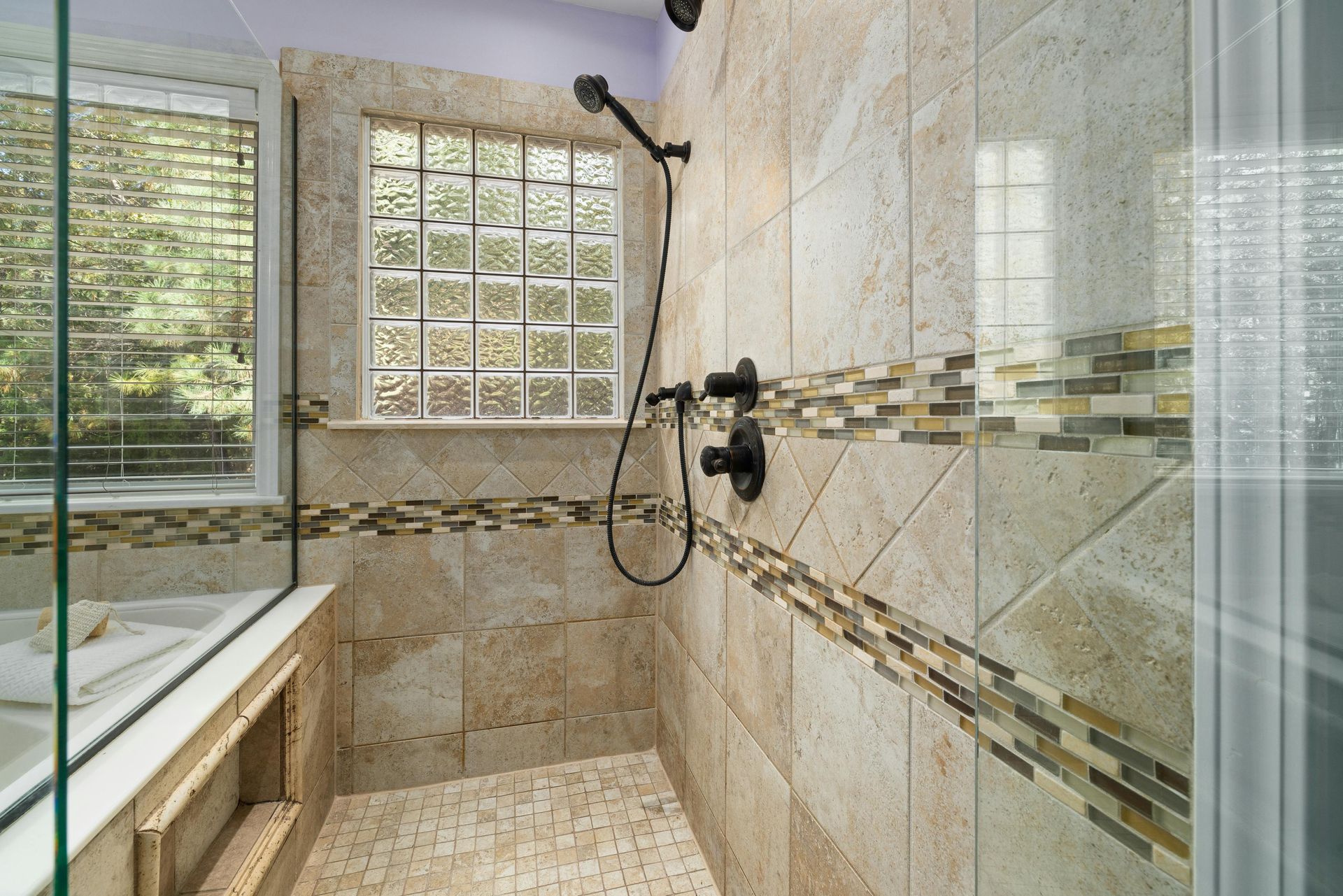 A walk-in shower with beige stone-look tiles, a decorative mosaic stripe, a handheld sprayer, and a glass block window.