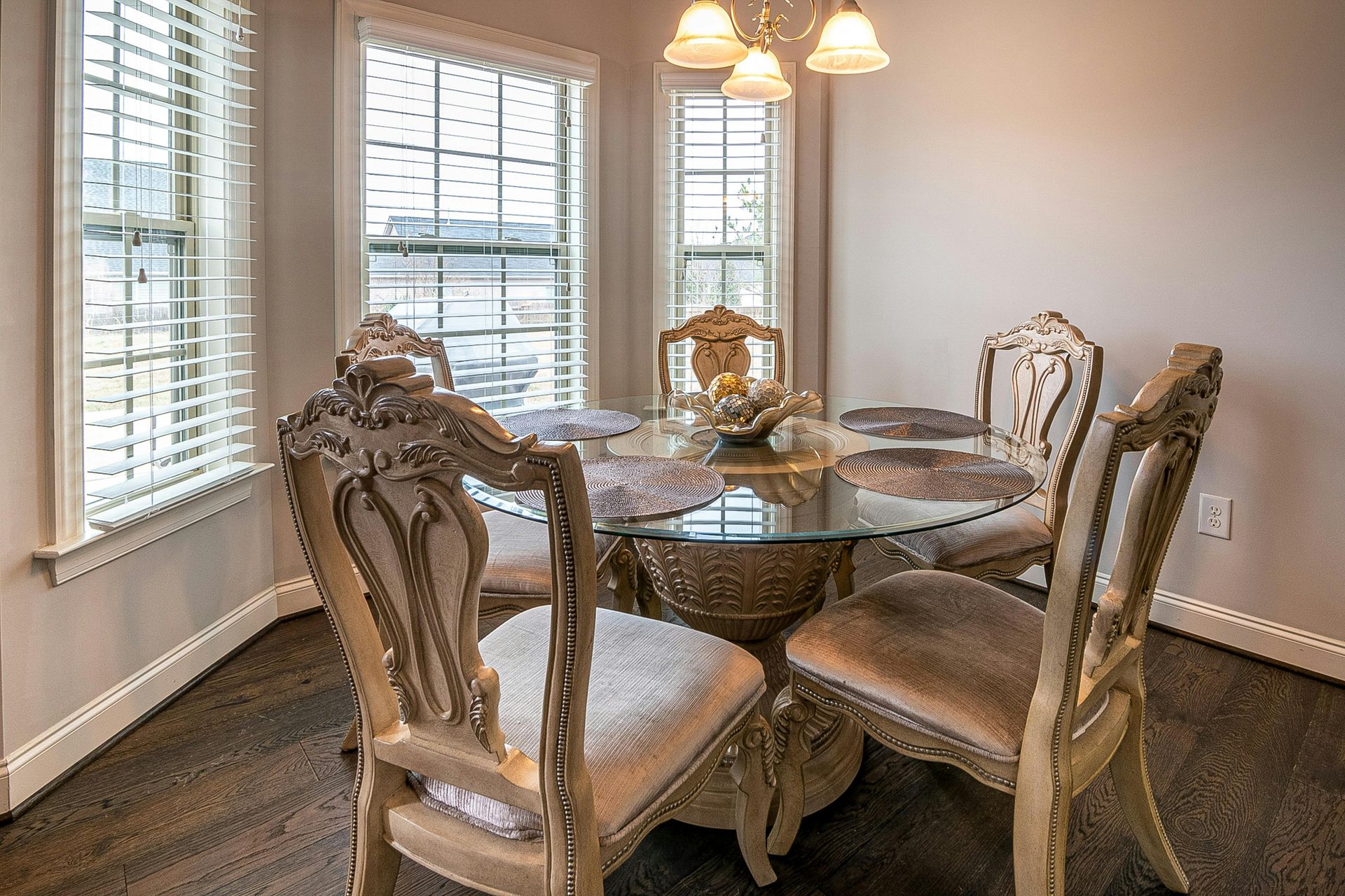 A dining set with a round glass table and four ornate, light-colored wooden chairs in a sunlit breakfast nook.
