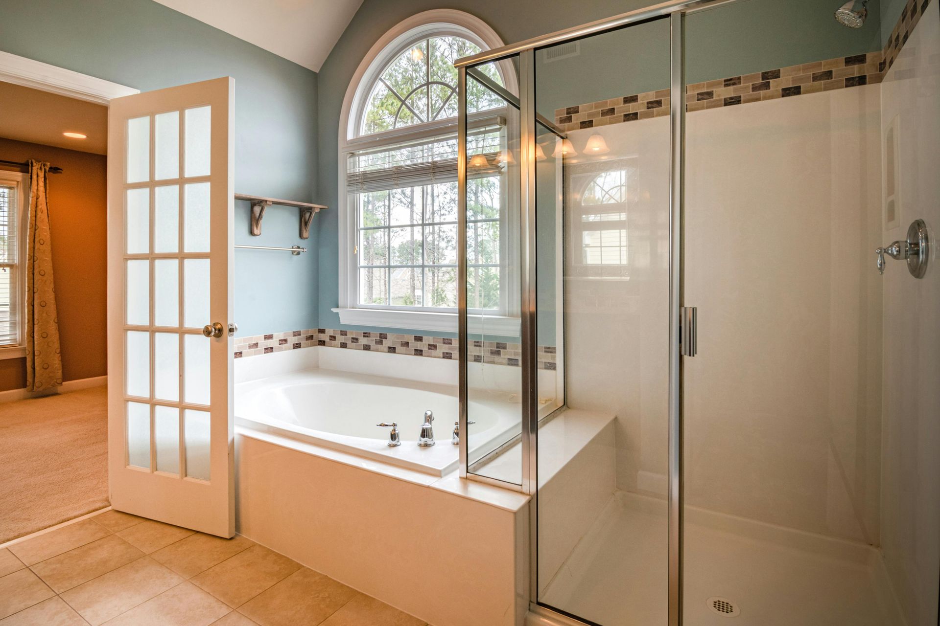 A bright bathroom features a soaking tub below an arched window, a glass-enclosed shower, and a French door to a bedroom.