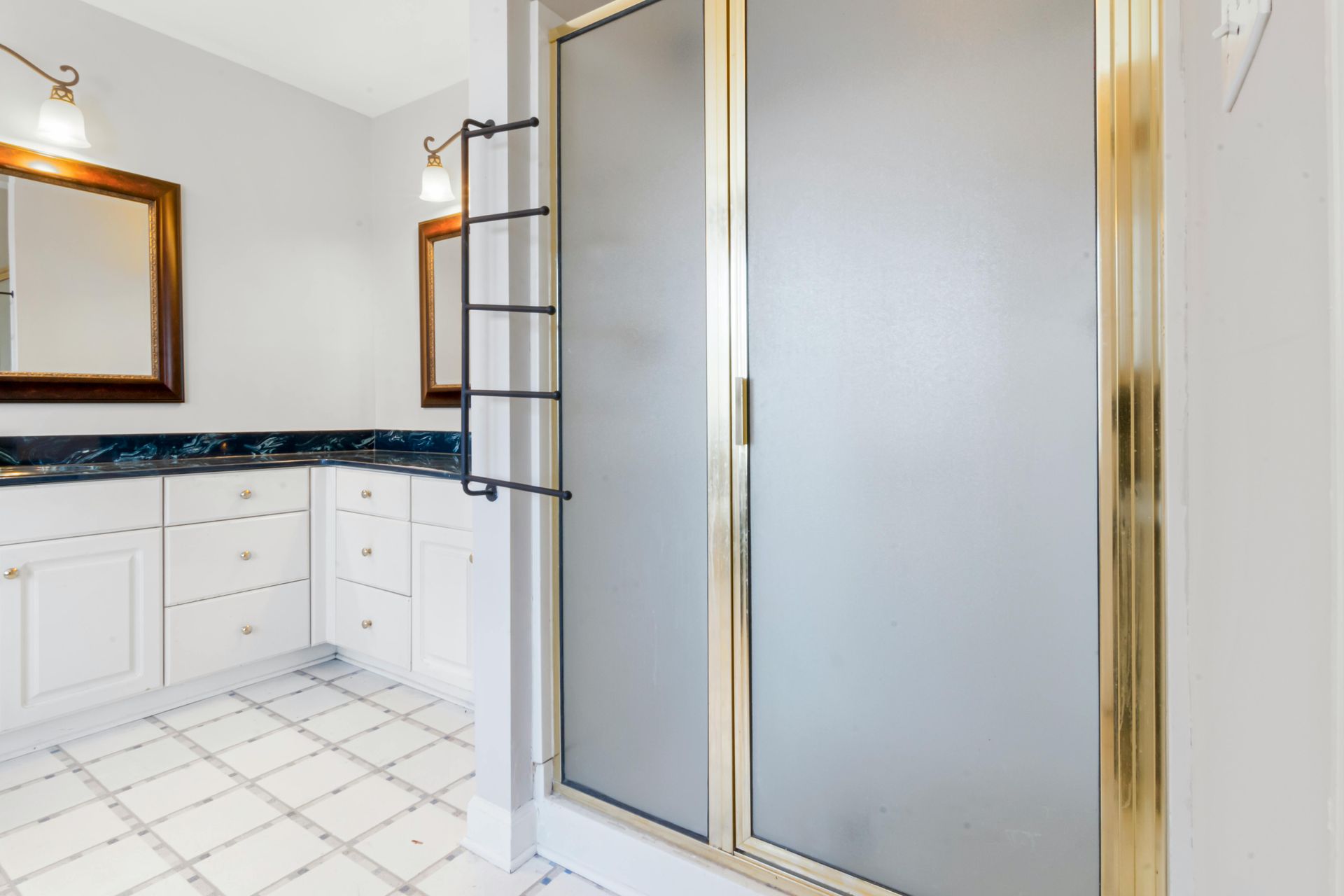 A brightly lit bathroom with a white vanity, dark countertops, tiled floors, and a glass-enclosed shower with gold trim.