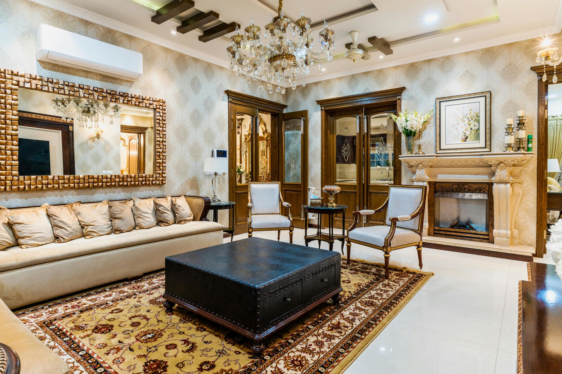 Luxurious living room featuring a long beige sofa, dark leather ottoman, ornate chandelier, and fireplace.