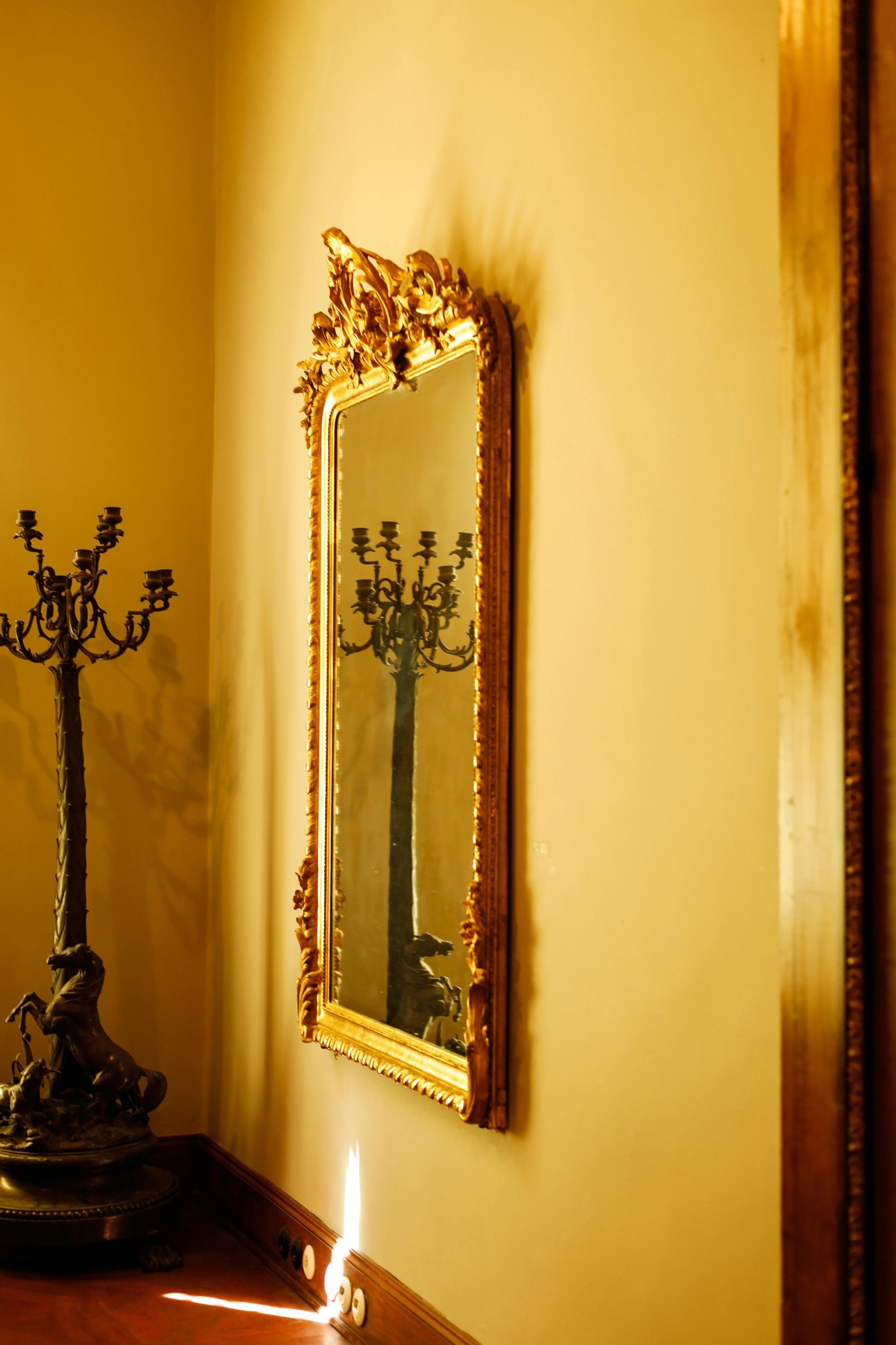 An ornate gold-framed wall mirror reflects a dark, multi-armed candle holder standing on a floor with bright sunlight.