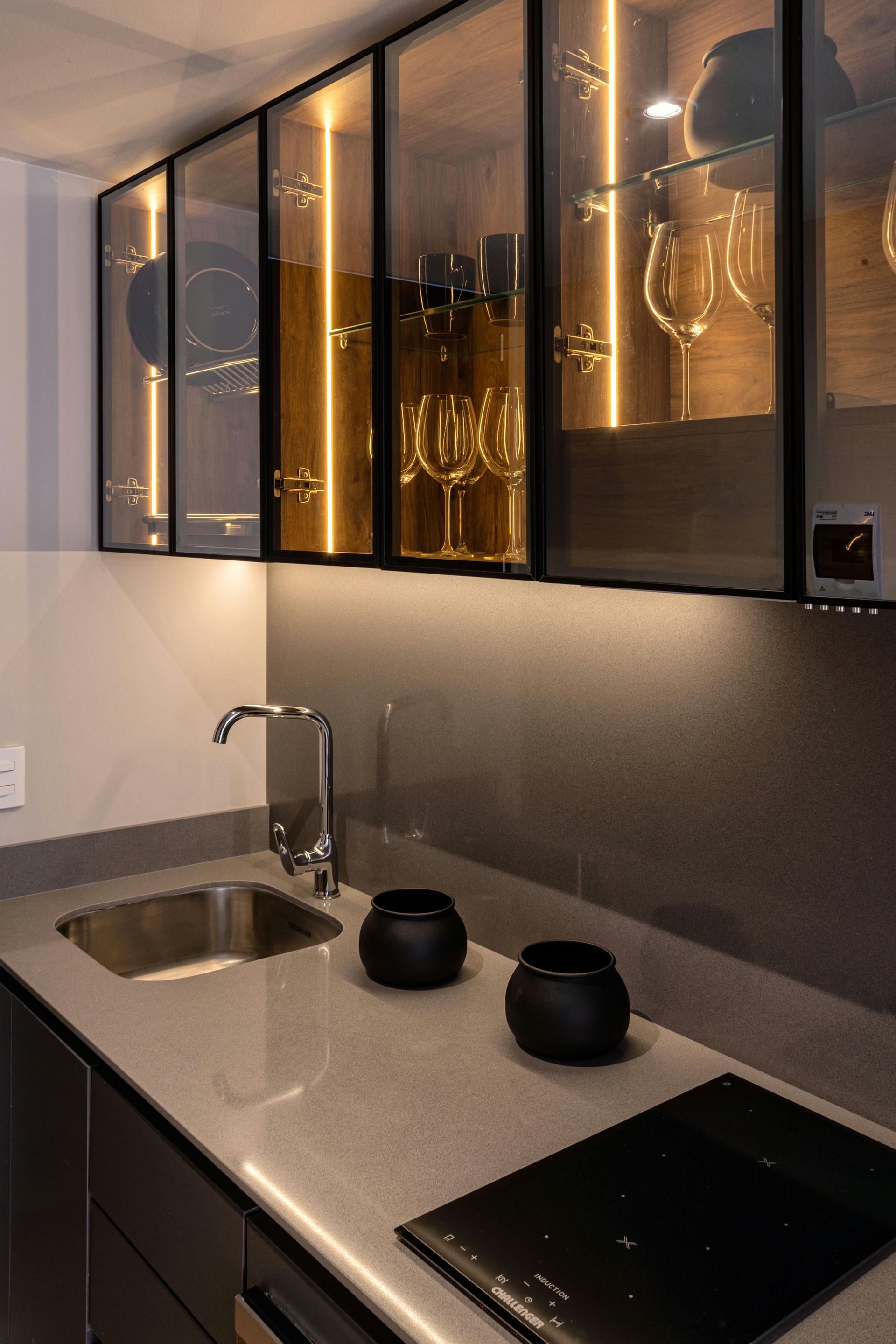 A modern kitchen with a grey countertop, sink, black cooktop, two black bowls, and illuminated glass-front cabinets.