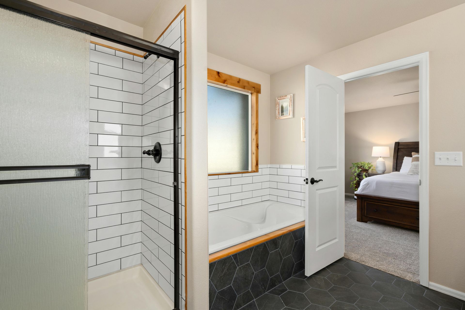 A bathroom featuring a glass-enclosed shower, a tub with dark tile surround, and an open door leading to a bedroom.