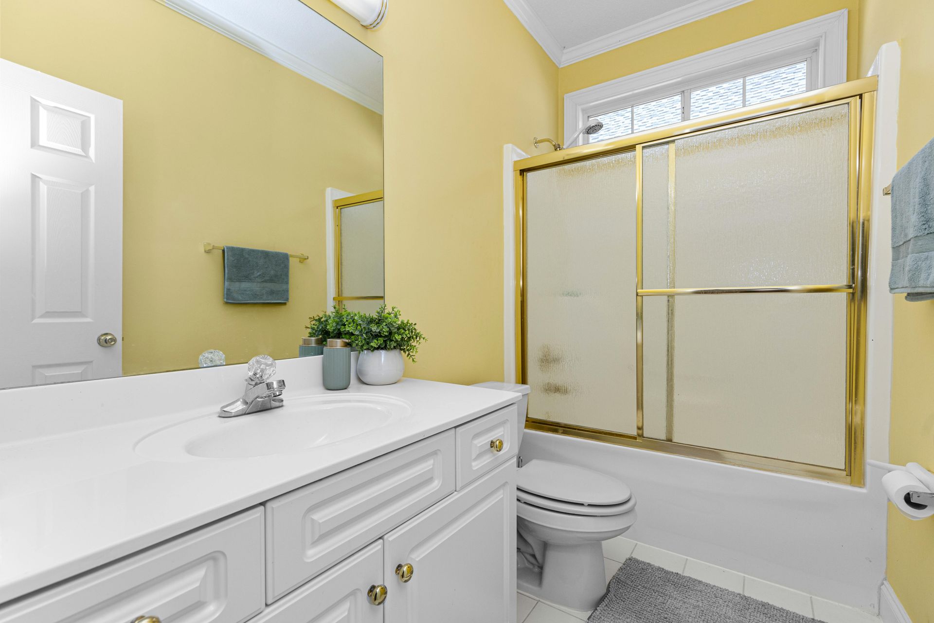 A bright yellow bathroom features a white vanity, a large mirror, a toilet, and a glass-enclosed shower with gold trim.
