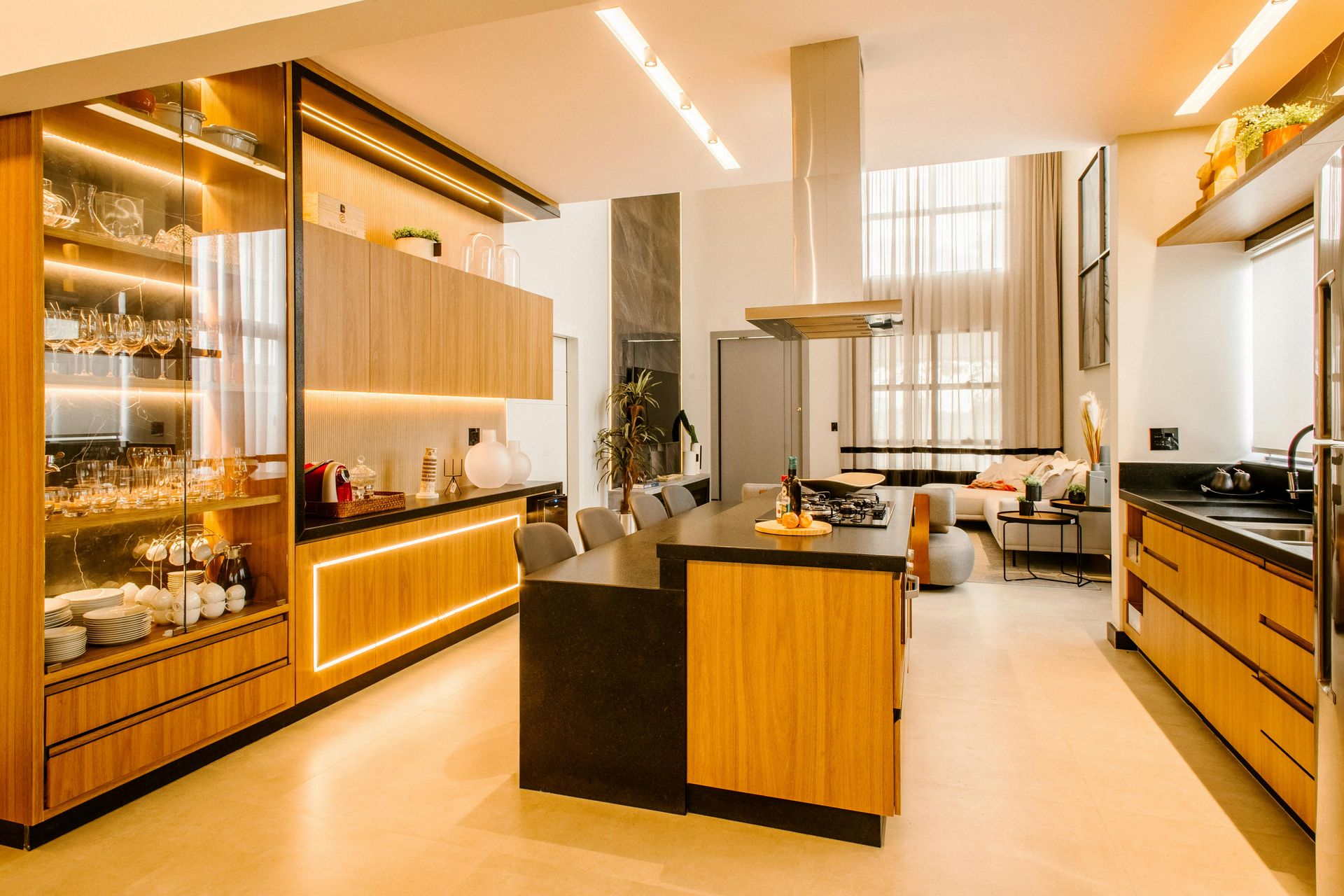 A modern, well-lit kitchen with light wood cabinetry, a large central island, black countertops, and glass display cases.