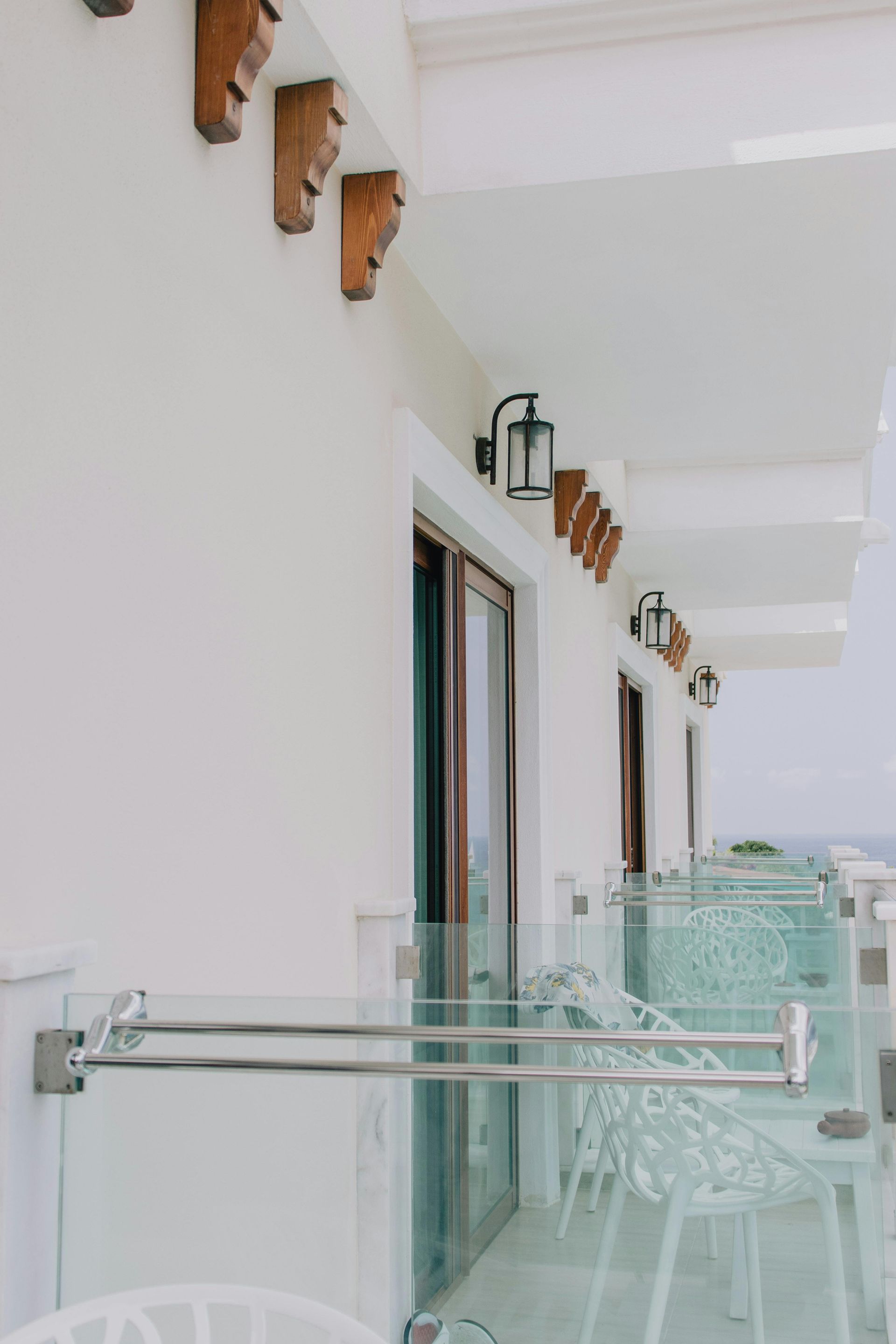 A row of balconies with glass railings, white walls, decorative wooden corbels, and outdoor chairs overlooking the sea.