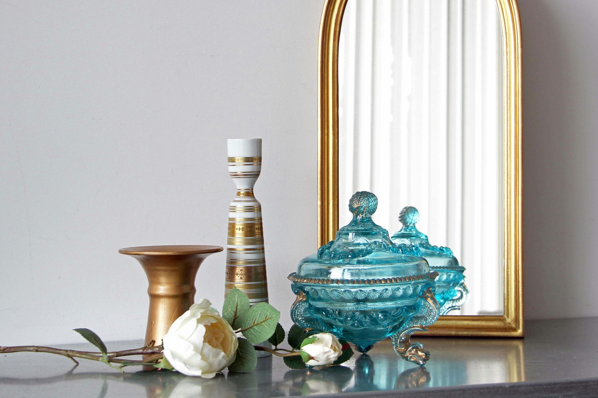 A decorative display with a brass candle holder, a tall patterned vase, blue glass jar, and white flowers by a mirror.