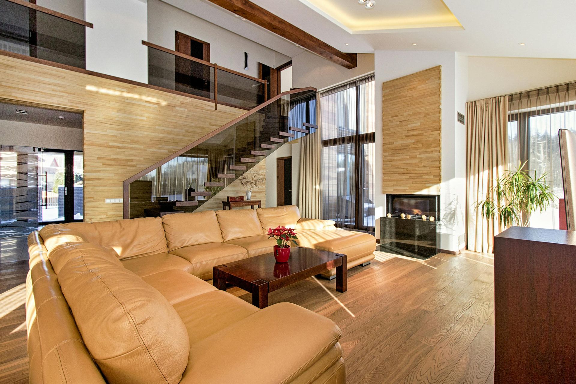 Modern living room featuring a tan leather sectional sofa, a dark wood coffee table, a glass-railed staircase, and fireplace.