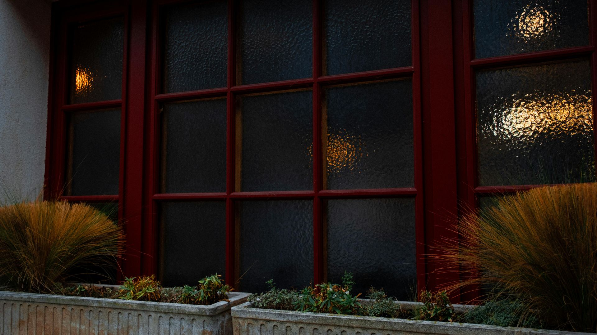 A row of frosted, red-framed window panes above outdoor stone planters filled with grasses and small green plants.