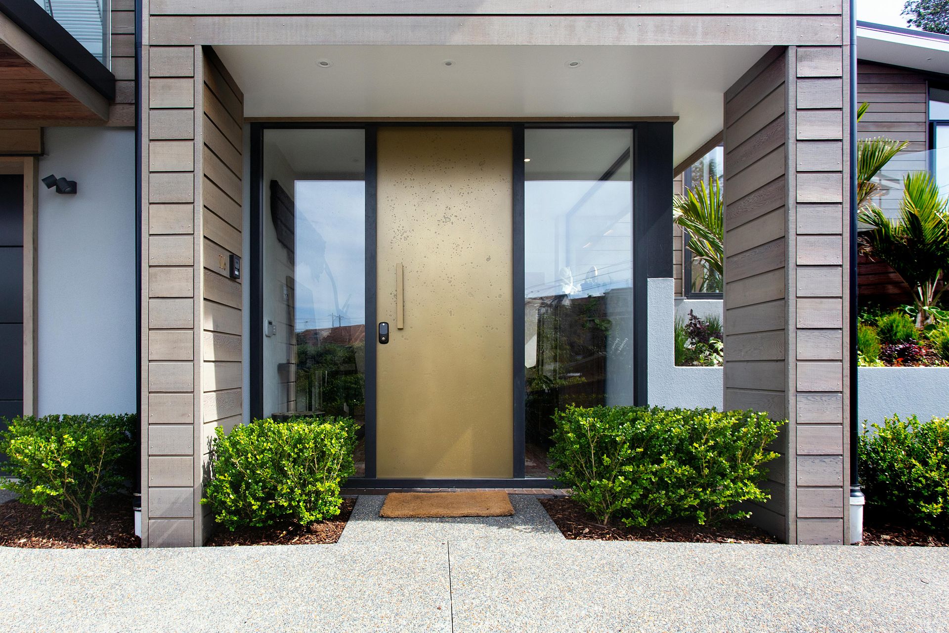 A modern entryway featuring a gold-toned door between two glass panels, framed by horizontal wood siding and low bushes.