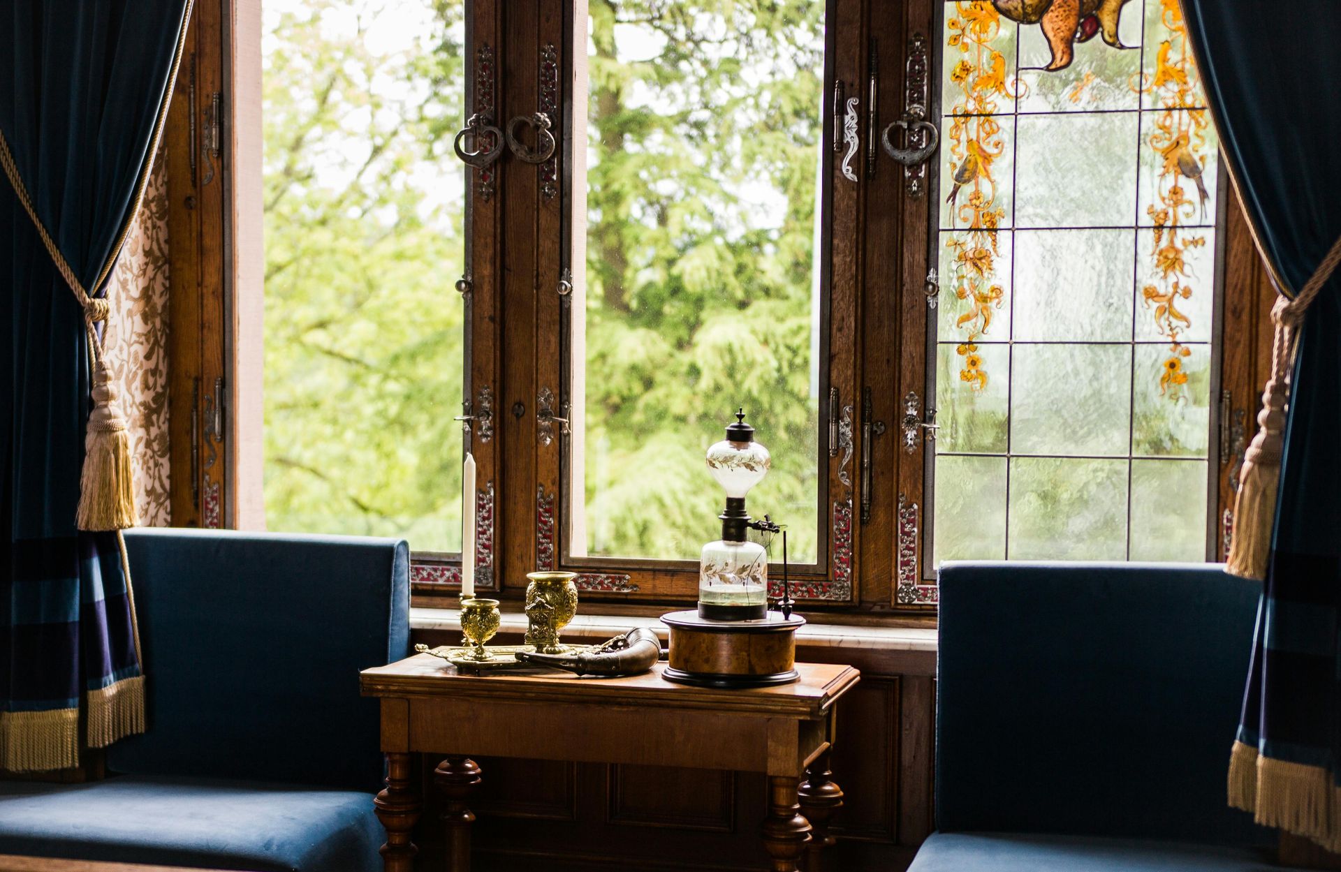 A small wooden table holds a candle and a lamp between two blue chairs in front of an ornate window with green views.
