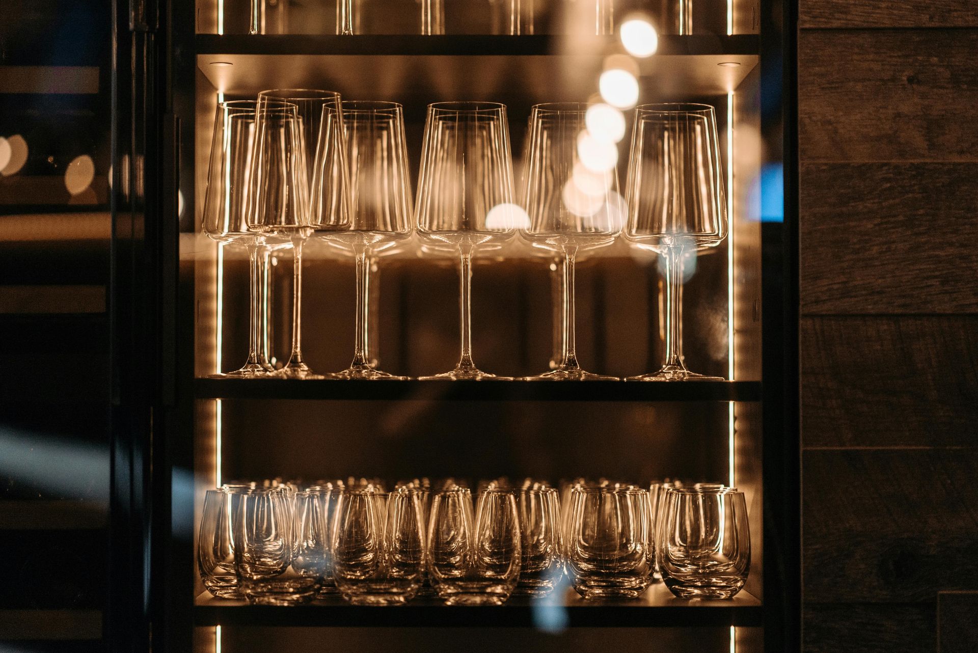 Two rows of clean, empty wine and water glasses displayed inside a modern, warm-lit glass cabinet.