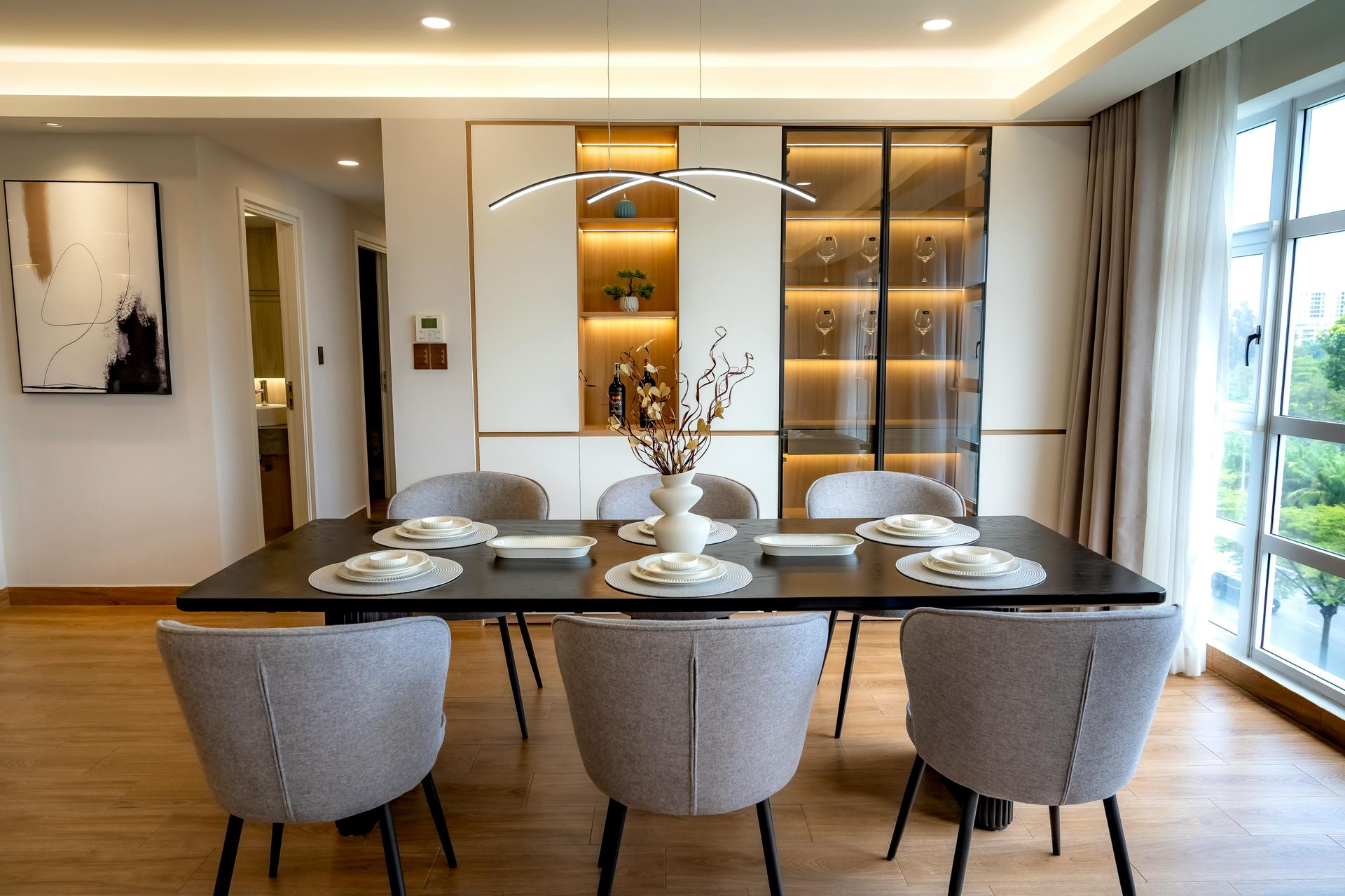 A modern dining room featuring a dark rectangular table with six gray upholstered chairs in front of a lit display unit.