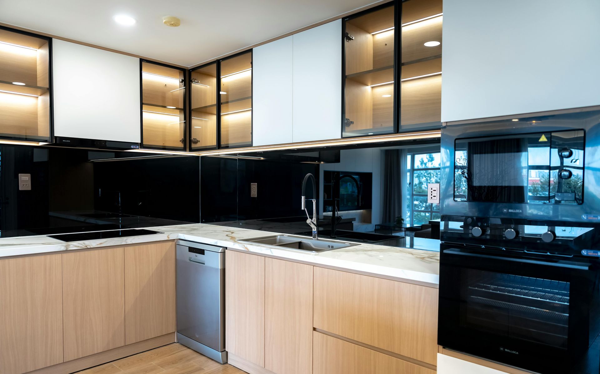 A modern kitchen with light wood cabinets, white upper cabinets, lit glass-front cupboards, a dishwasher, and a wall oven.