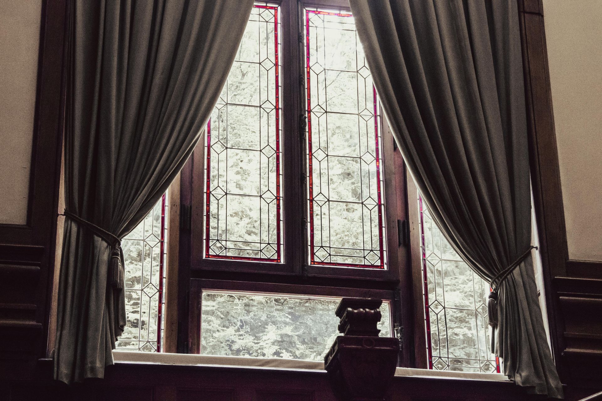 A window with leaded glass panes framed by long, draped sage-green curtains in a wood-paneled room.