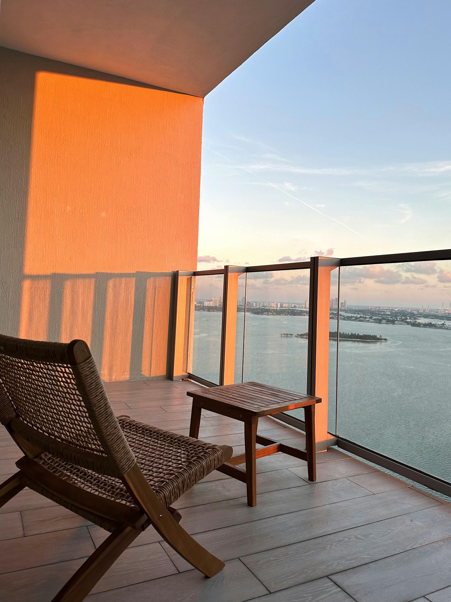 A wooden chair and small table on a balcony overlooking a bay at sunset, with golden light hitting a textured wall.