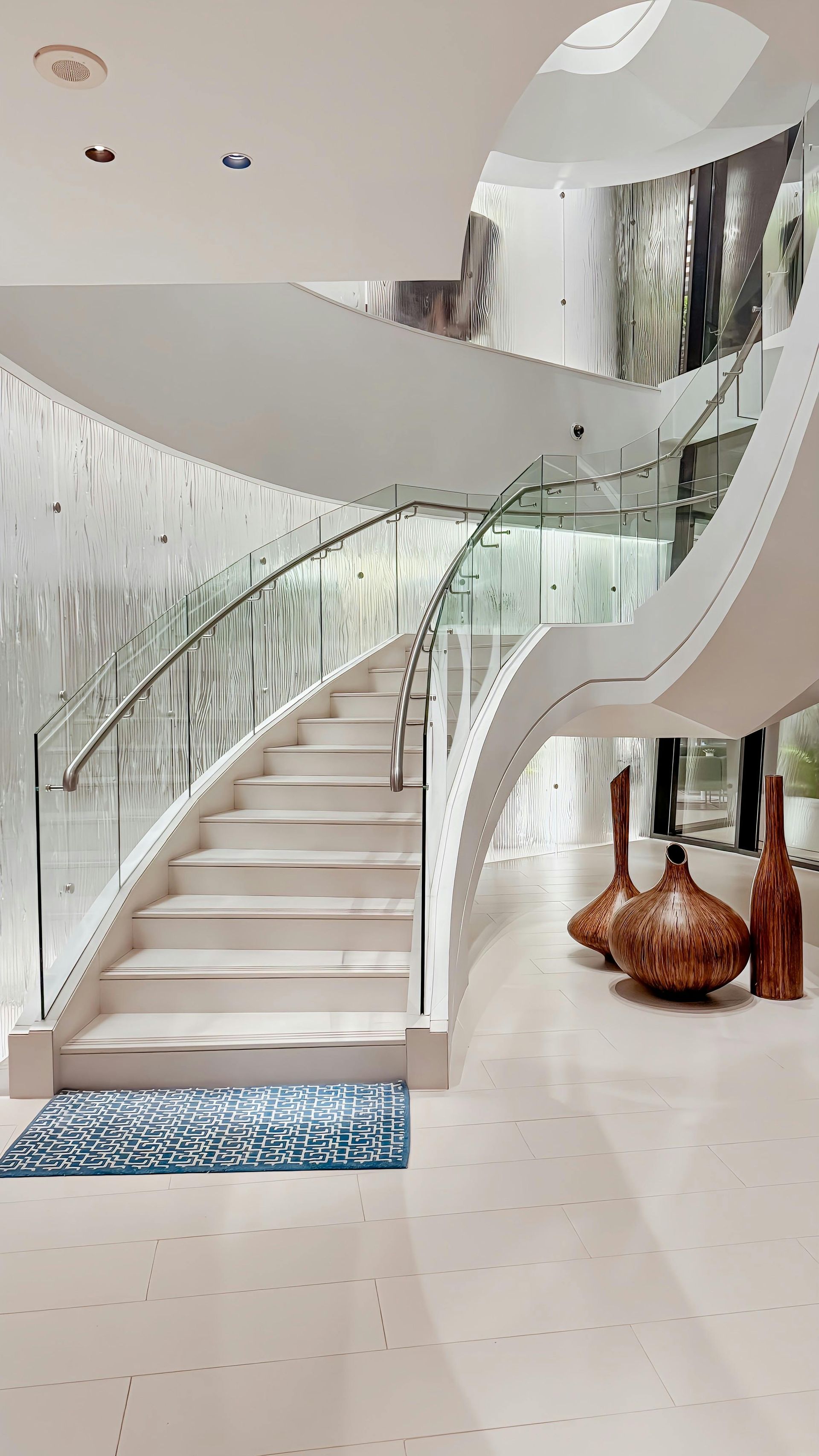A white, curved staircase with a glass railing stands in a modern, bright lobby next to sculptural bronze vessels.