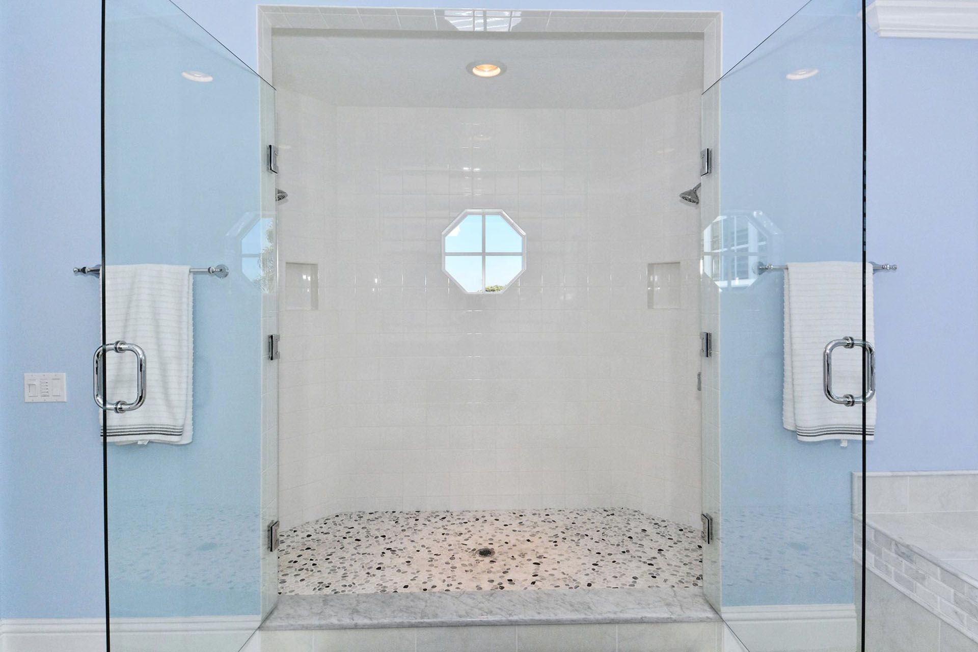 Glass shower enclosure with blue walls and a hexagonal window above a pebble-stone shower floor