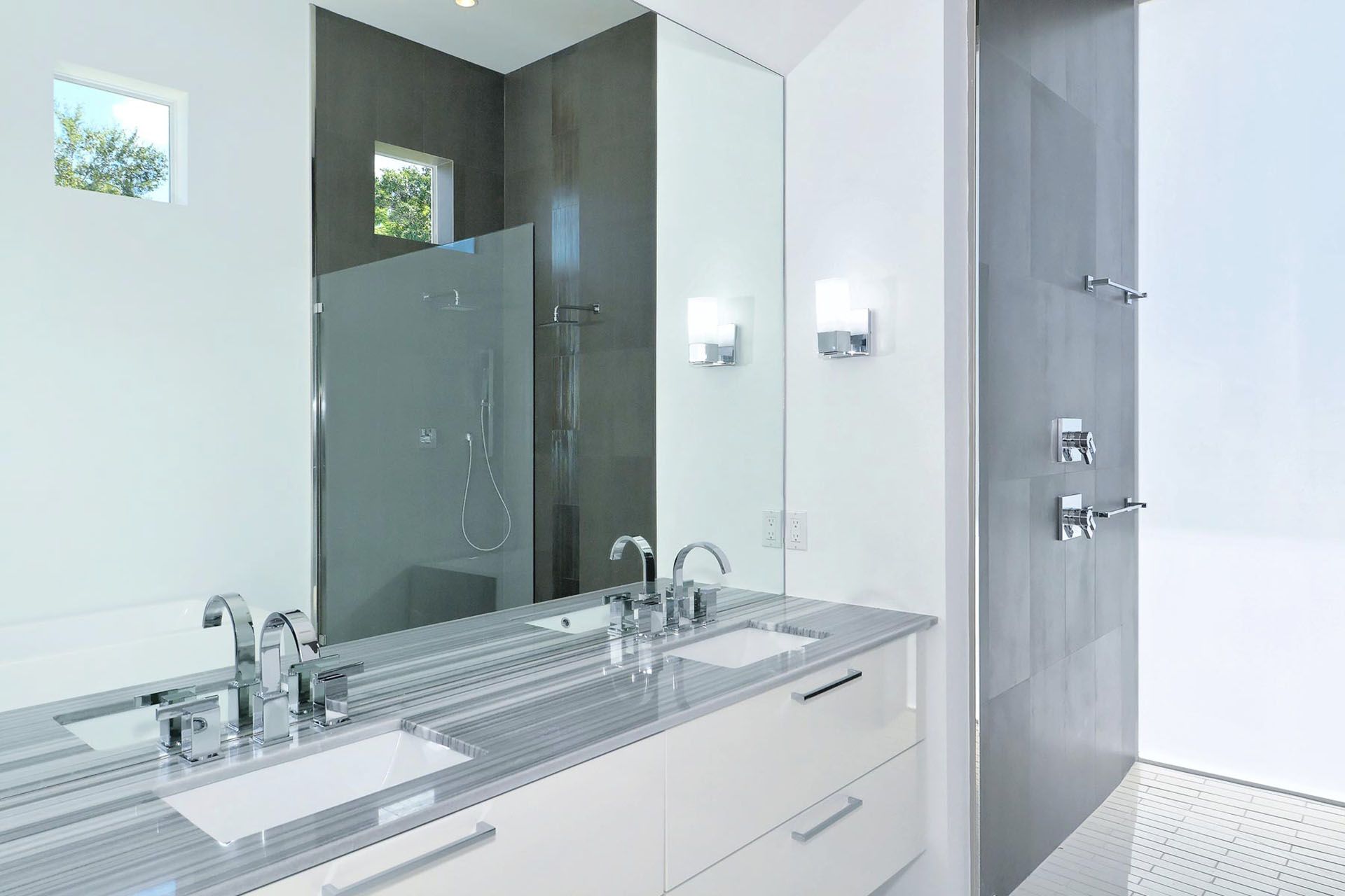 Modern bathroom vanity with double sinks, large mirror, glass shower, and gray tile walls