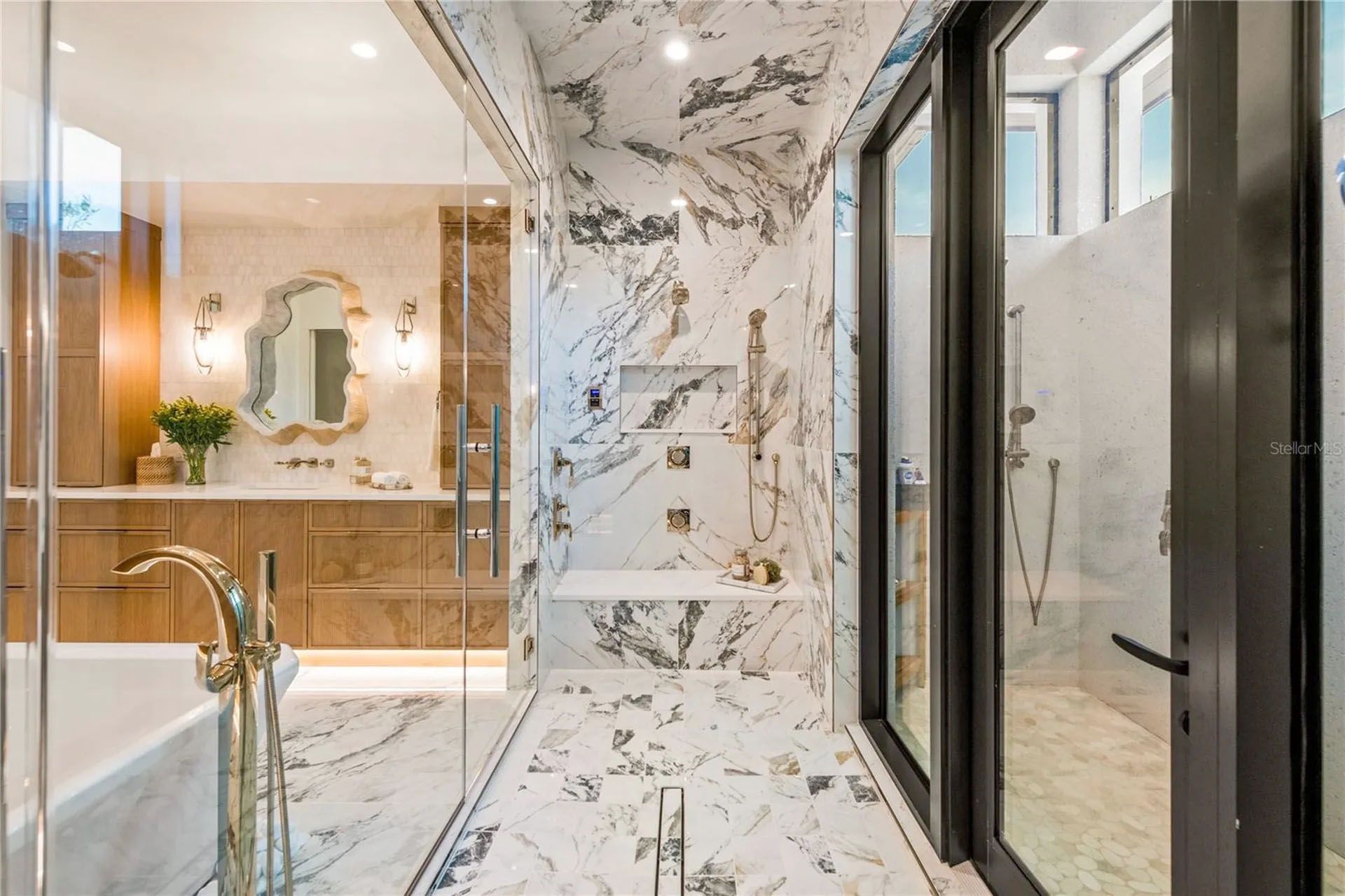 Modern bathroom with marble walls, glass shower, double vanity, and large mirror reflective of the bright interior