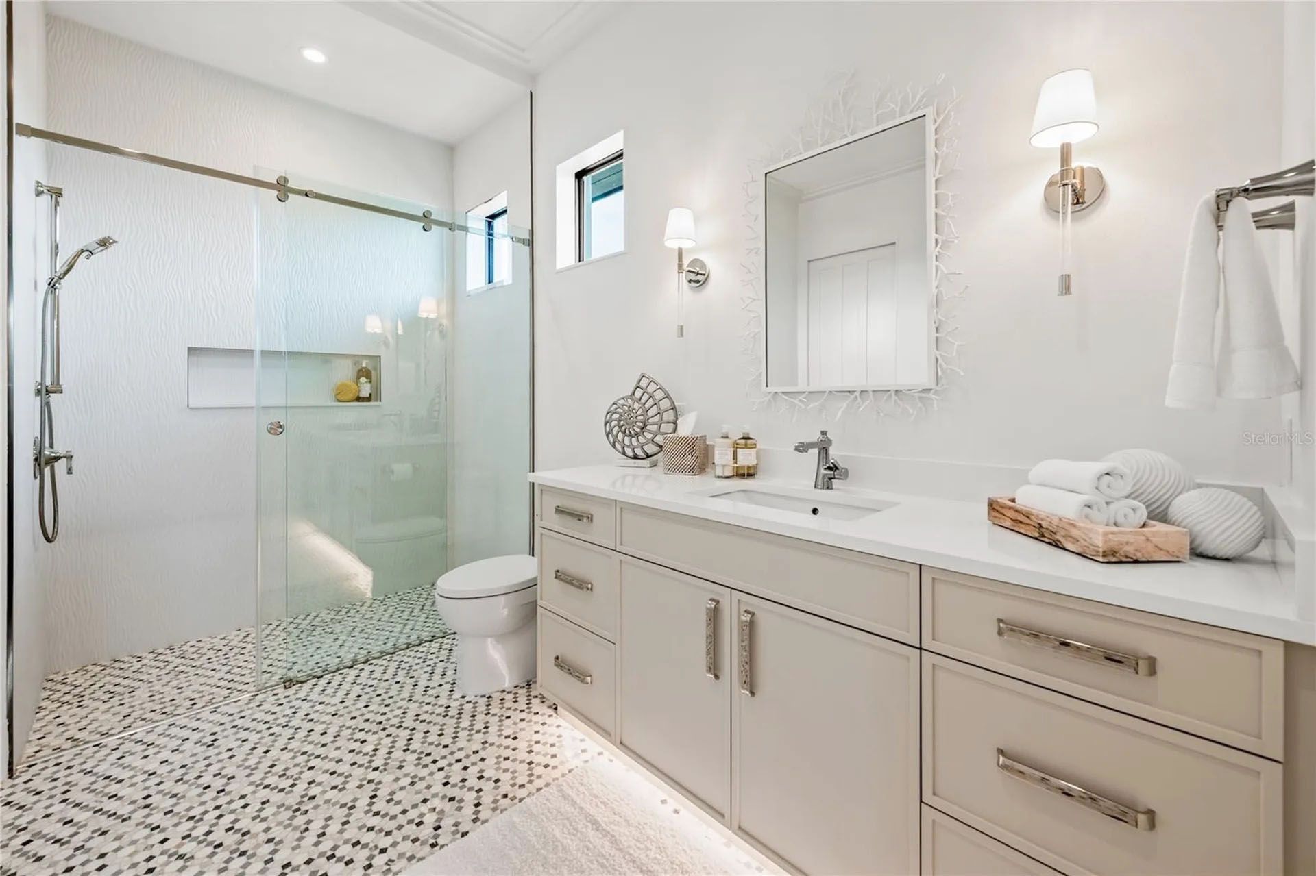 Bright modern bathroom with glass shower, double vanity, white walls, and patterned tile floor