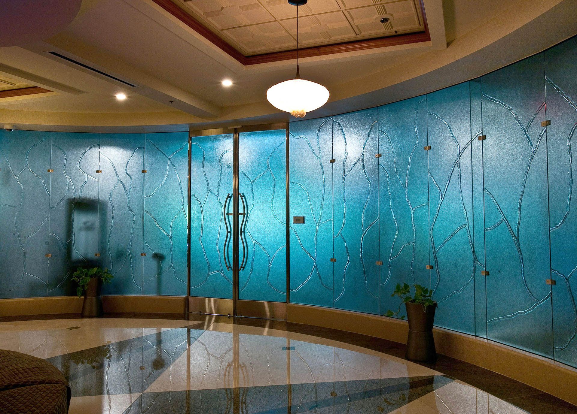Elegant lobby with blue textured glass wall, curved bench, potted plants, and central chandelier lighting