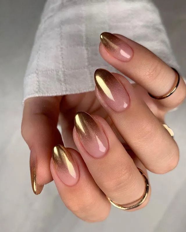 Golden ombre nails on a hand with gold rings, against a white long-sleeved shirt.