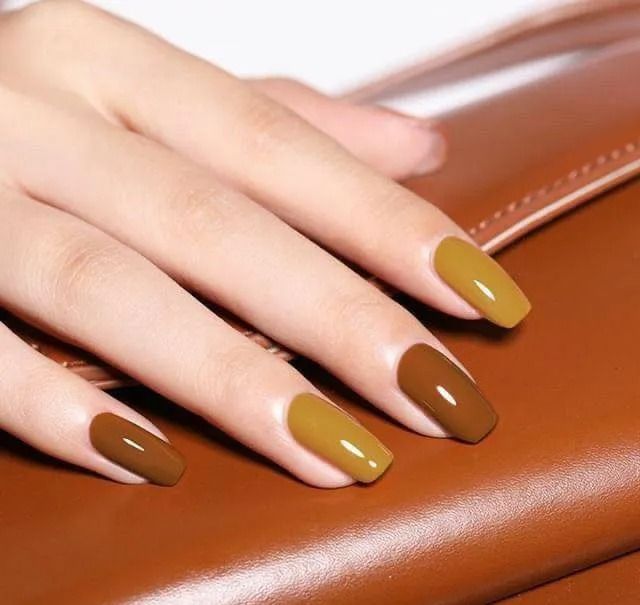 Hand with long, polished nails in shades of brown and mustard, resting on a brown surface.