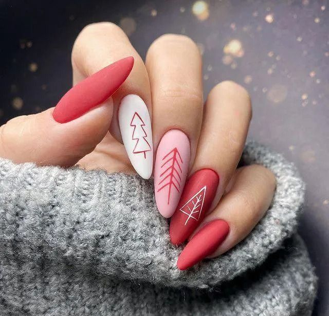 Hand with red, pink, and white matte Christmas-themed nail art featuring tree designs, on a grey sweater.