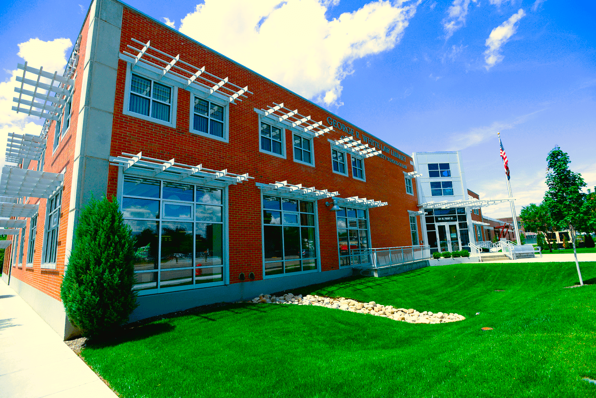 Two-story brick building with large windows, white pergolas, and a green lawn under a sunny, blue sky.