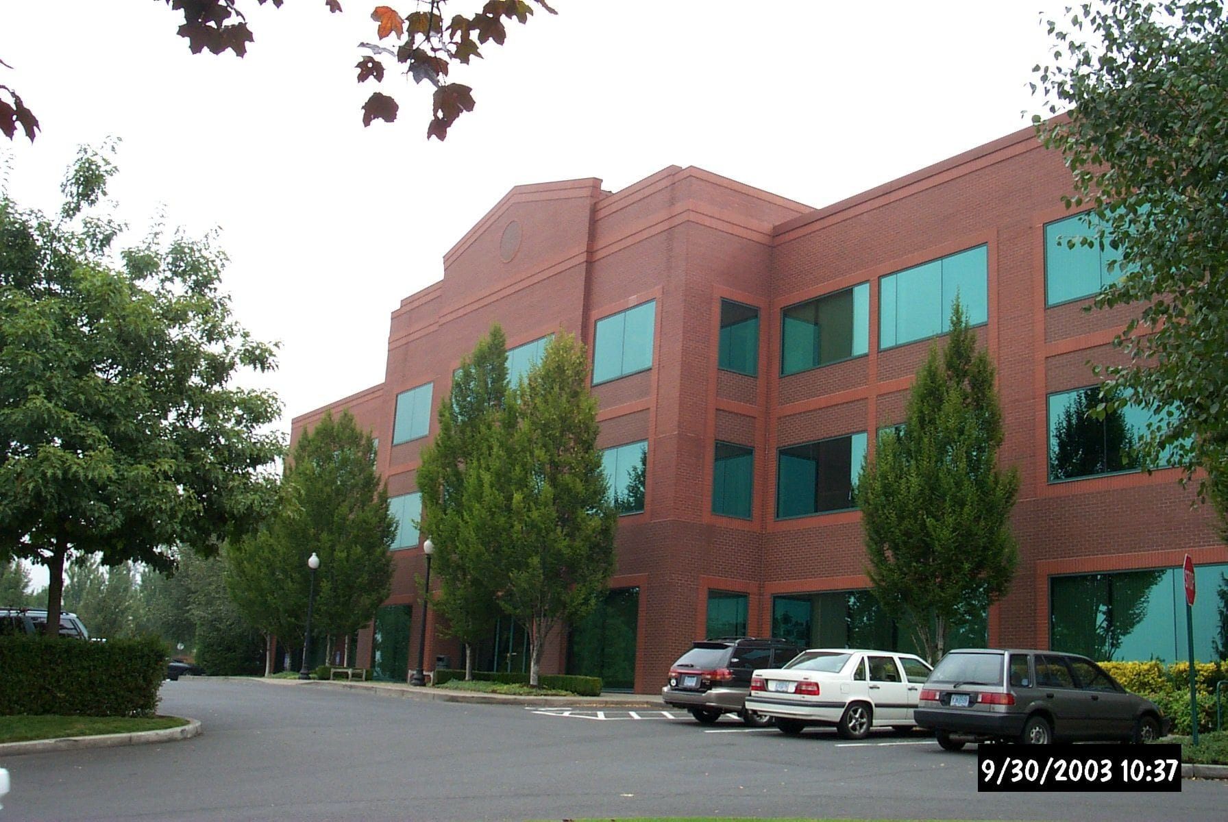 Red brick office building with teal-tinted windows, trees, and parked cars on a cloudy day.