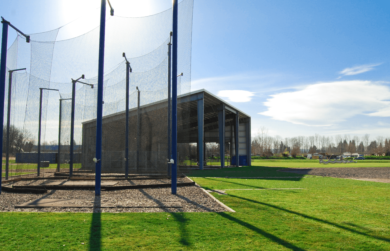 A baseball batting cage and field on a sunny day. A metallic building is behind the cage.
