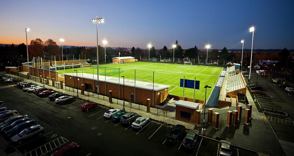 Football stadium at dusk with lights illuminating the field and parking lot full of cars.