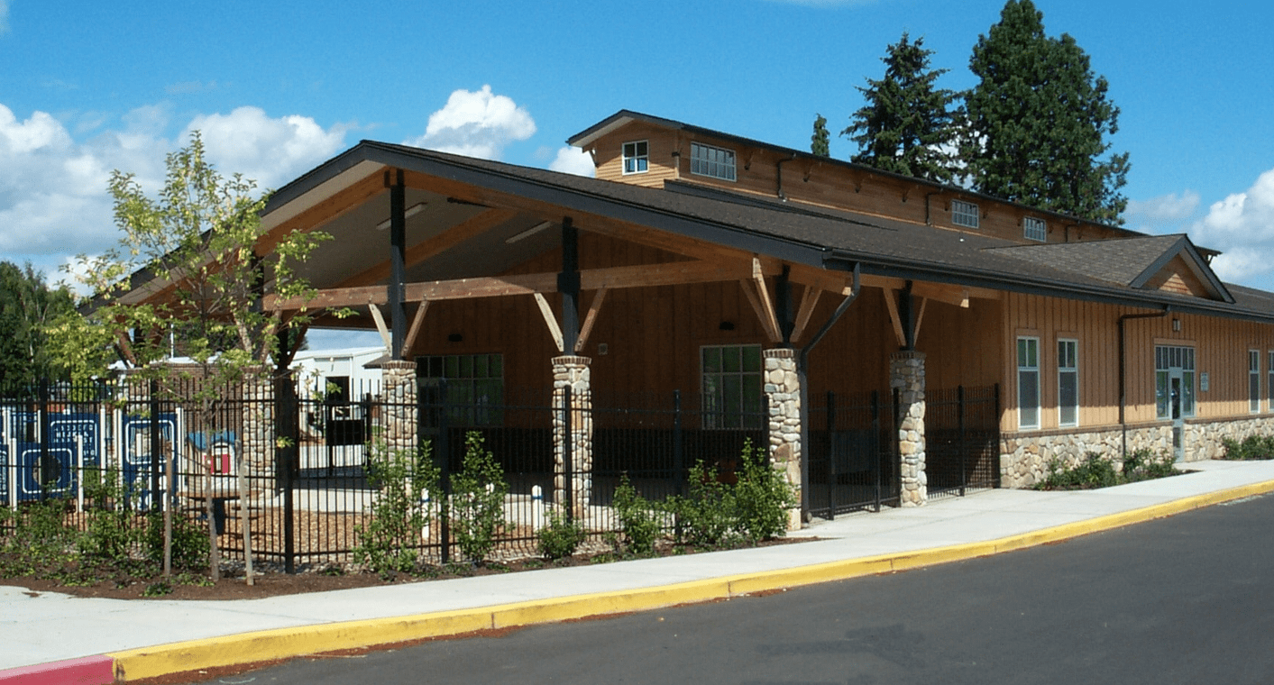 A wood-clad building with a covered entrance supported by stone columns.  A fence borders a sidewalk.