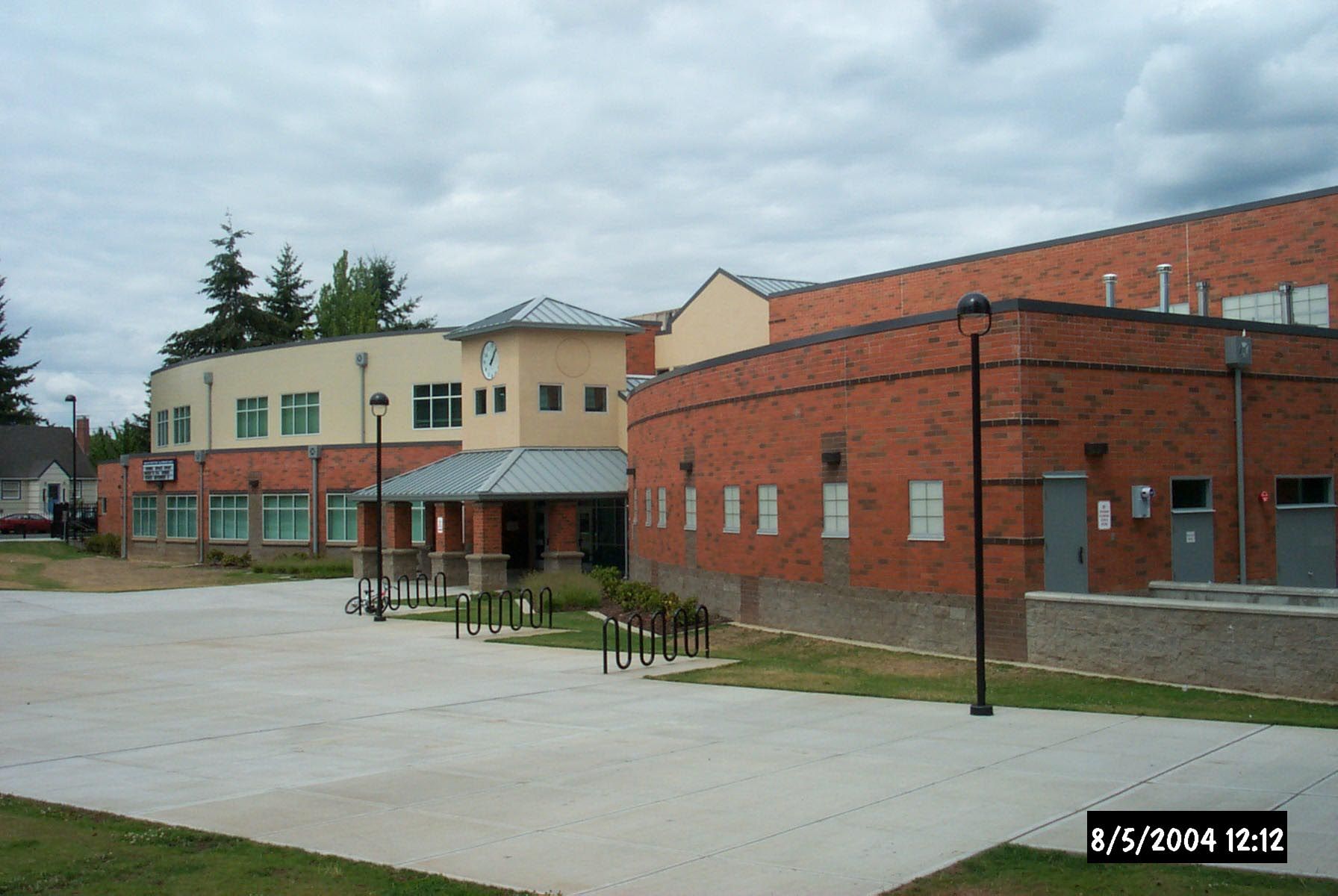 Exterior view of brick and light-colored buildings with a paved area and bike racks in front, under a cloudy sky.