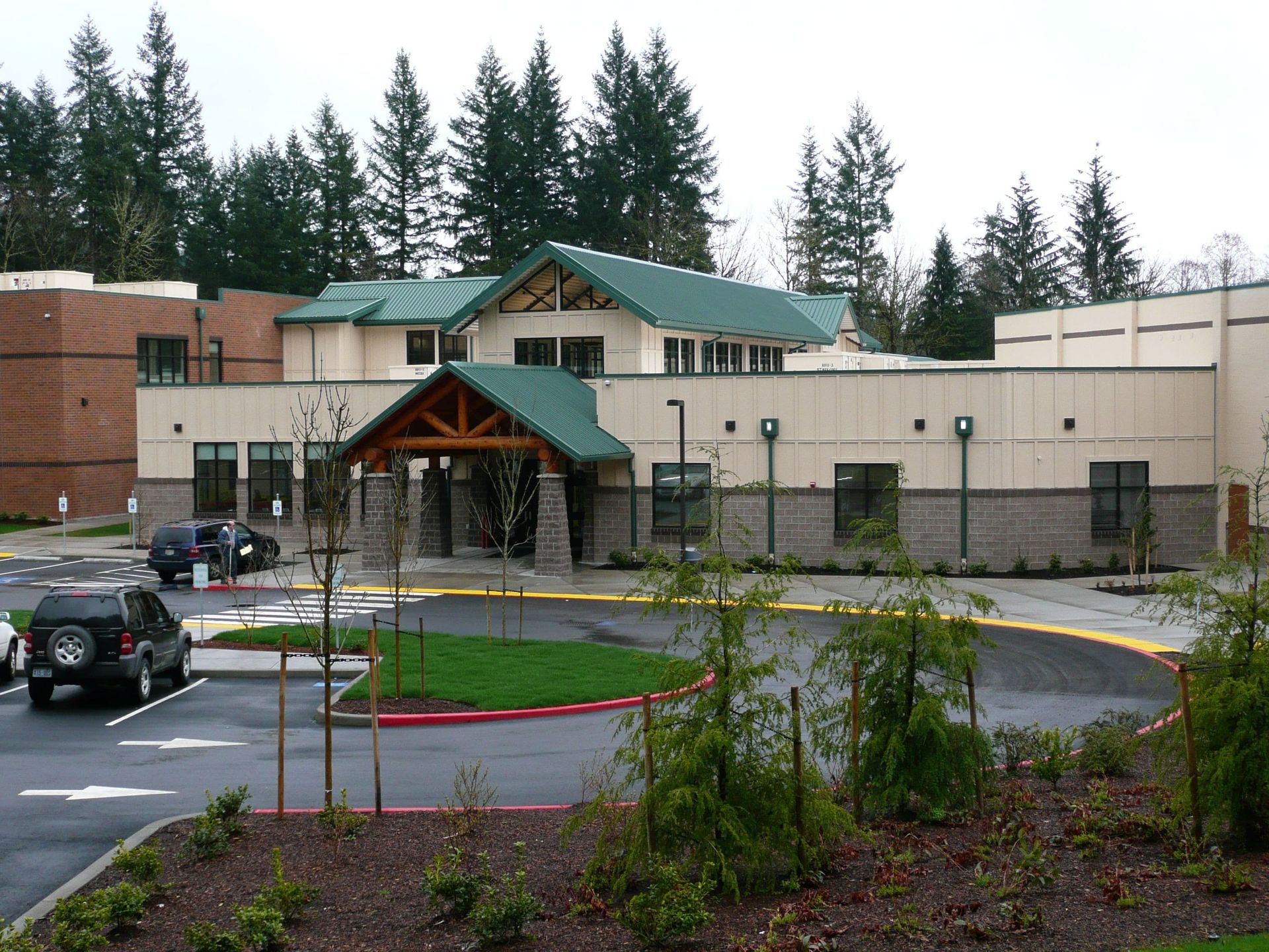 Modern school building with green roof, circular driveway, and several parked cars in front of a wooded backdrop.