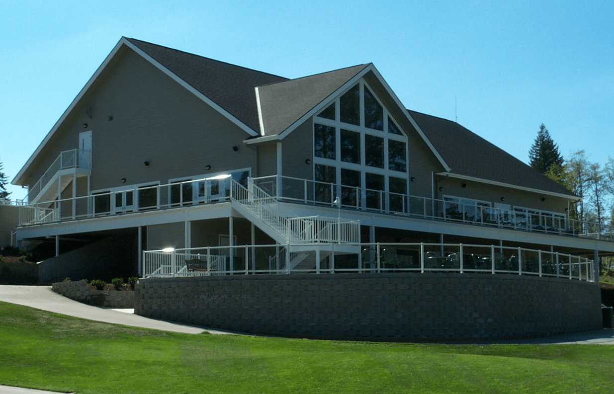 Tan building with large windows, wraparound balcony, and curved stone base. Green lawn in foreground.