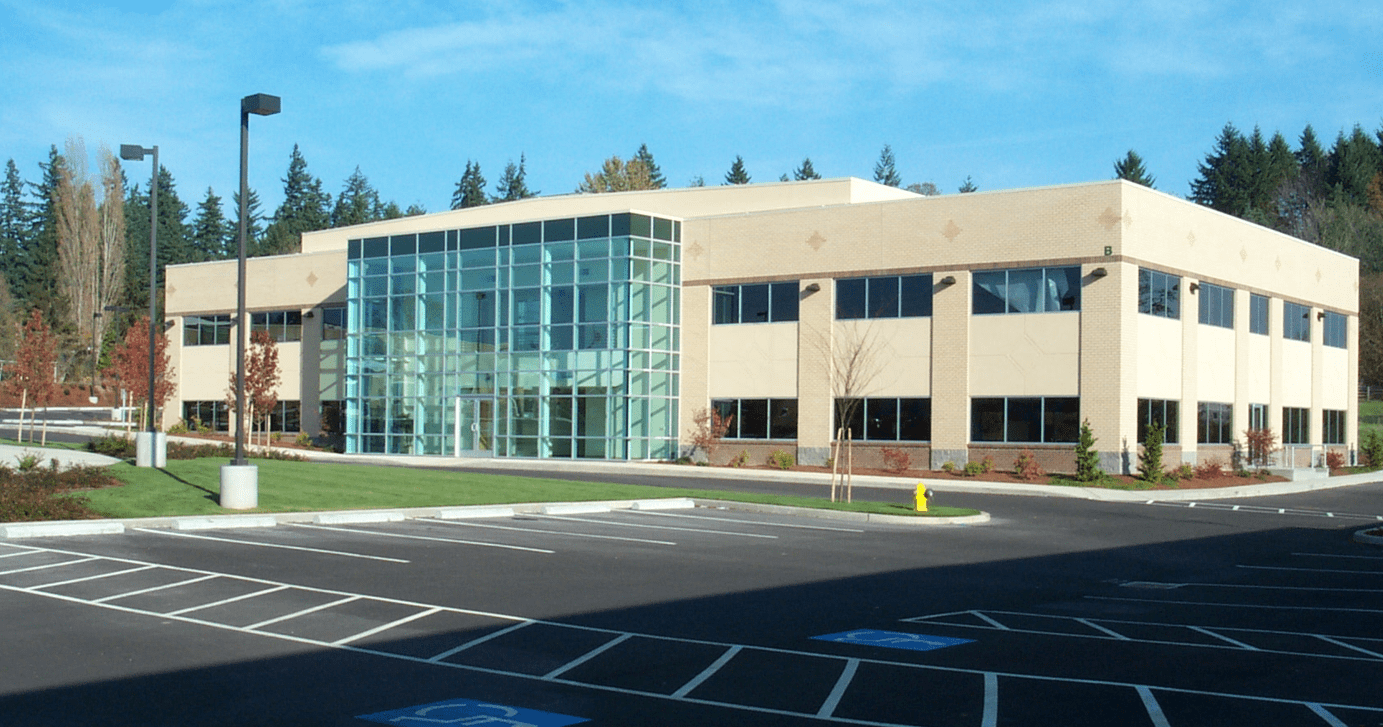 Modern tan office building with large glass entrance, surrounded by a parking lot and greenery.